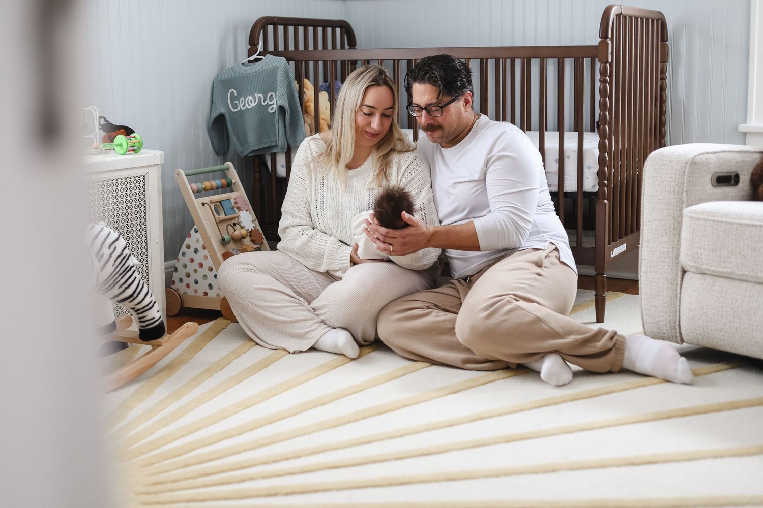 Parents sitting on nursery floor holding baby, with wooden crib and soft decor in background during in-home newborn session in Twin Cities