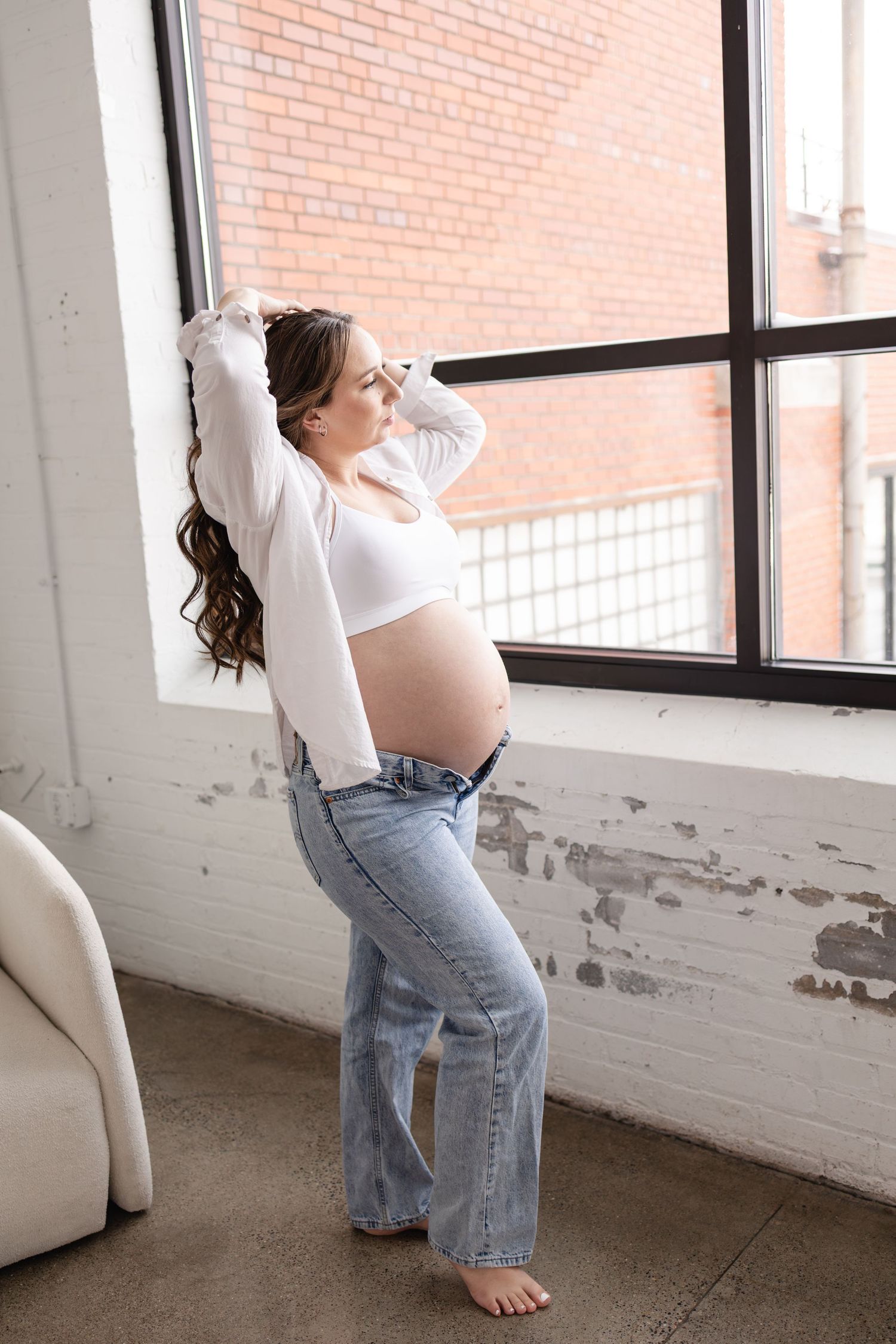 Pregnant woman in white crop top and jeans poses by large window, showing baby bump in bright loft space in Minneapolis