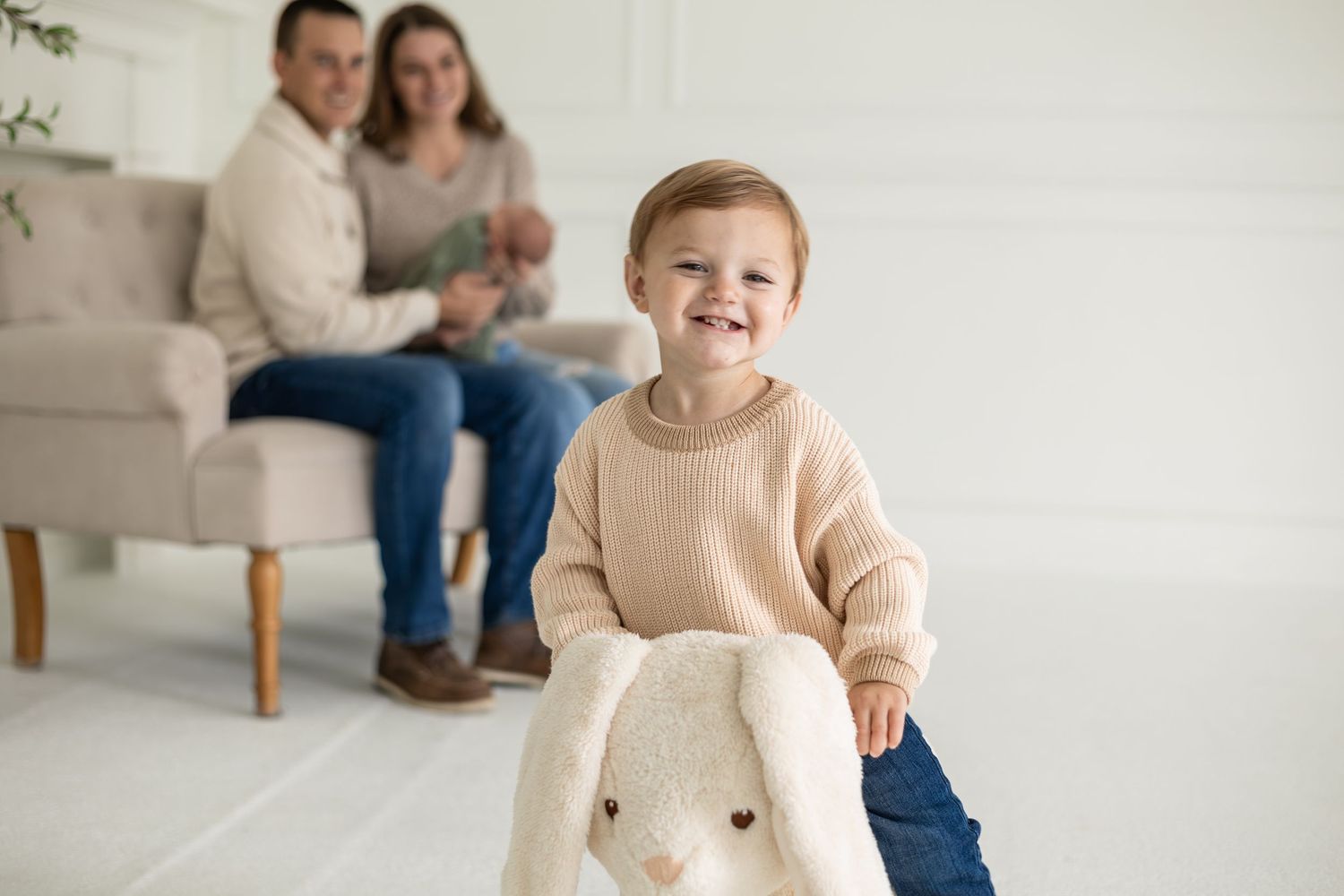 A small child in a beige sweater smiles while sitting on a rocking bunny during a newborn photo session in Twin Cities.