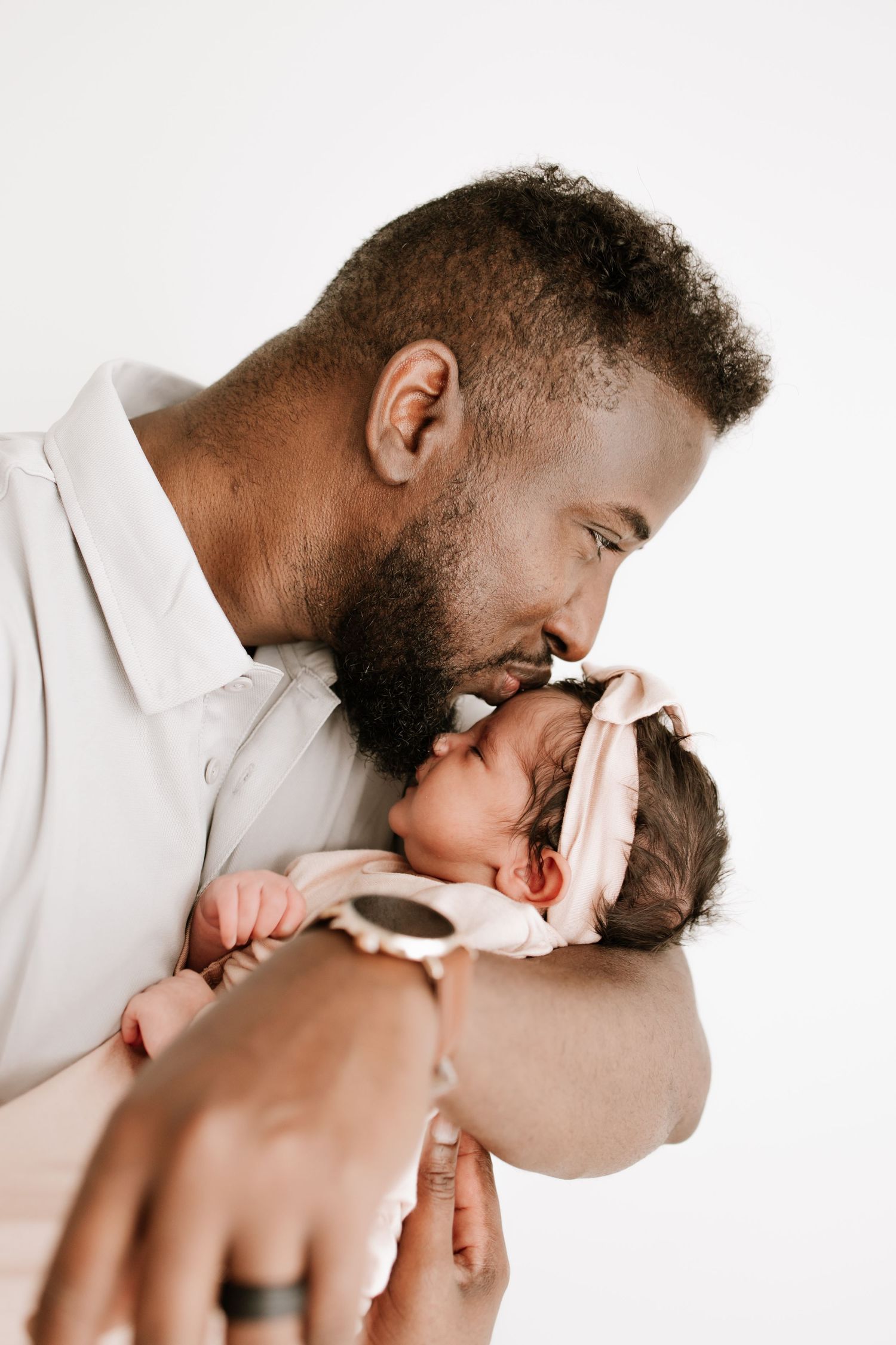 A loving father gently kisses his newborn baby girl wearing a pink headband in a bright studio in Farmington, MN.