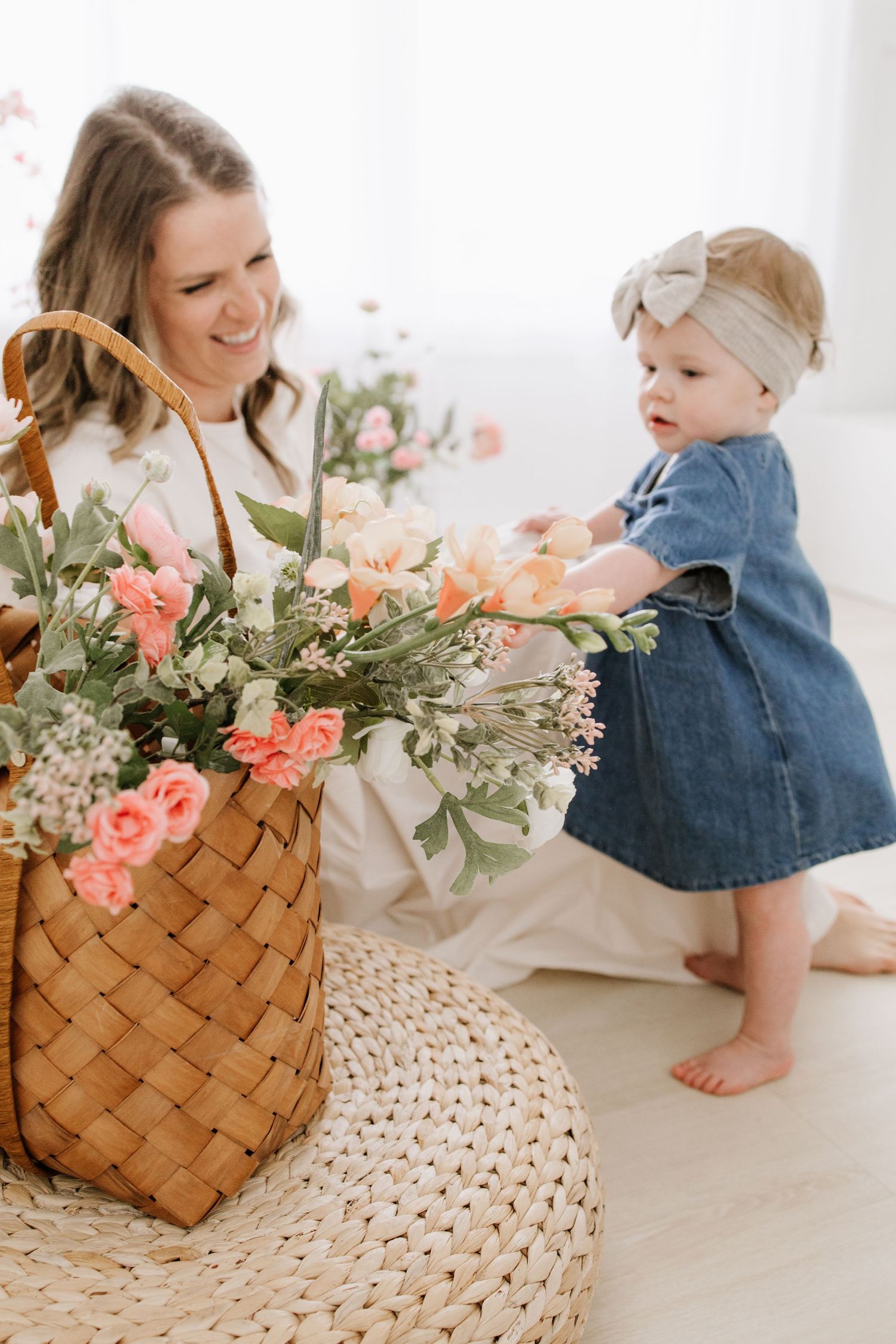A smiling mother watches her baby girl in a denim dress reach for pink flowers in a wicker basket in Lakeville studio mini session