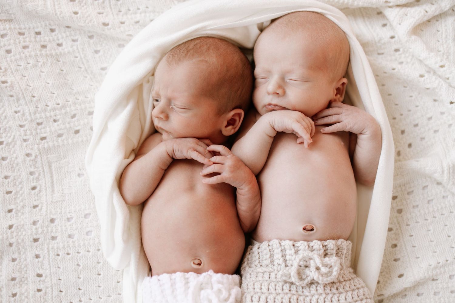 Two sleeping newborn twins wrapped in white knit blankets lying side by side in a cozy, soft white setting.