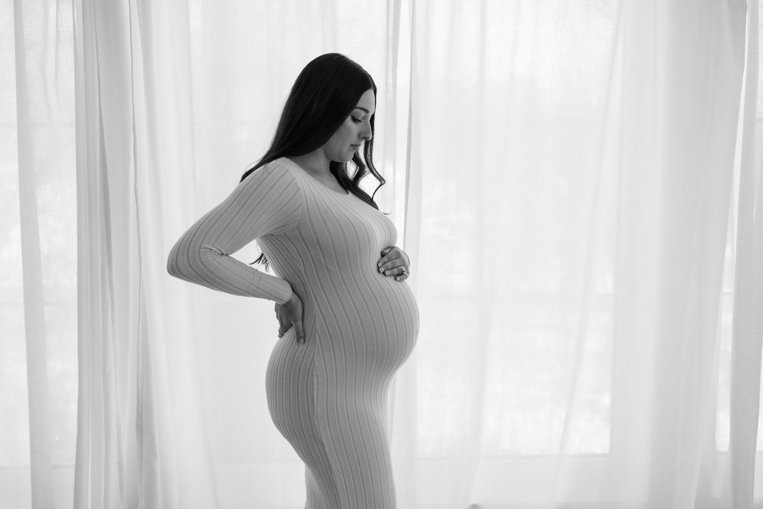 Silhouettes of a pregnant woman in white dress pose gracefully against a window with sheer curtains in a maternity photoshoot in Twin Cities