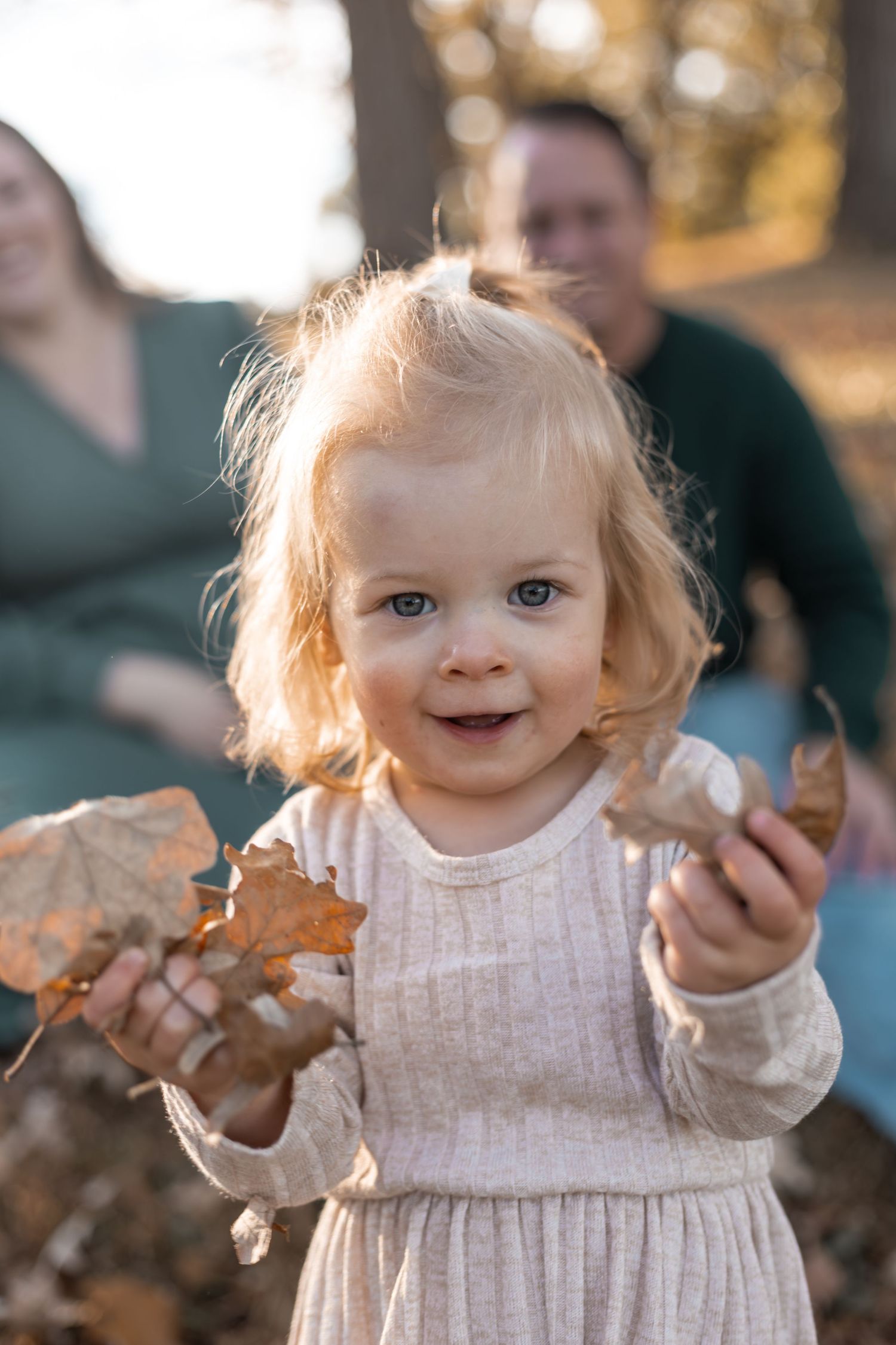Smiling toddler girl in cream dress holding autumn leaves outdoors with parents in background during fall family photo session in Lakeville.