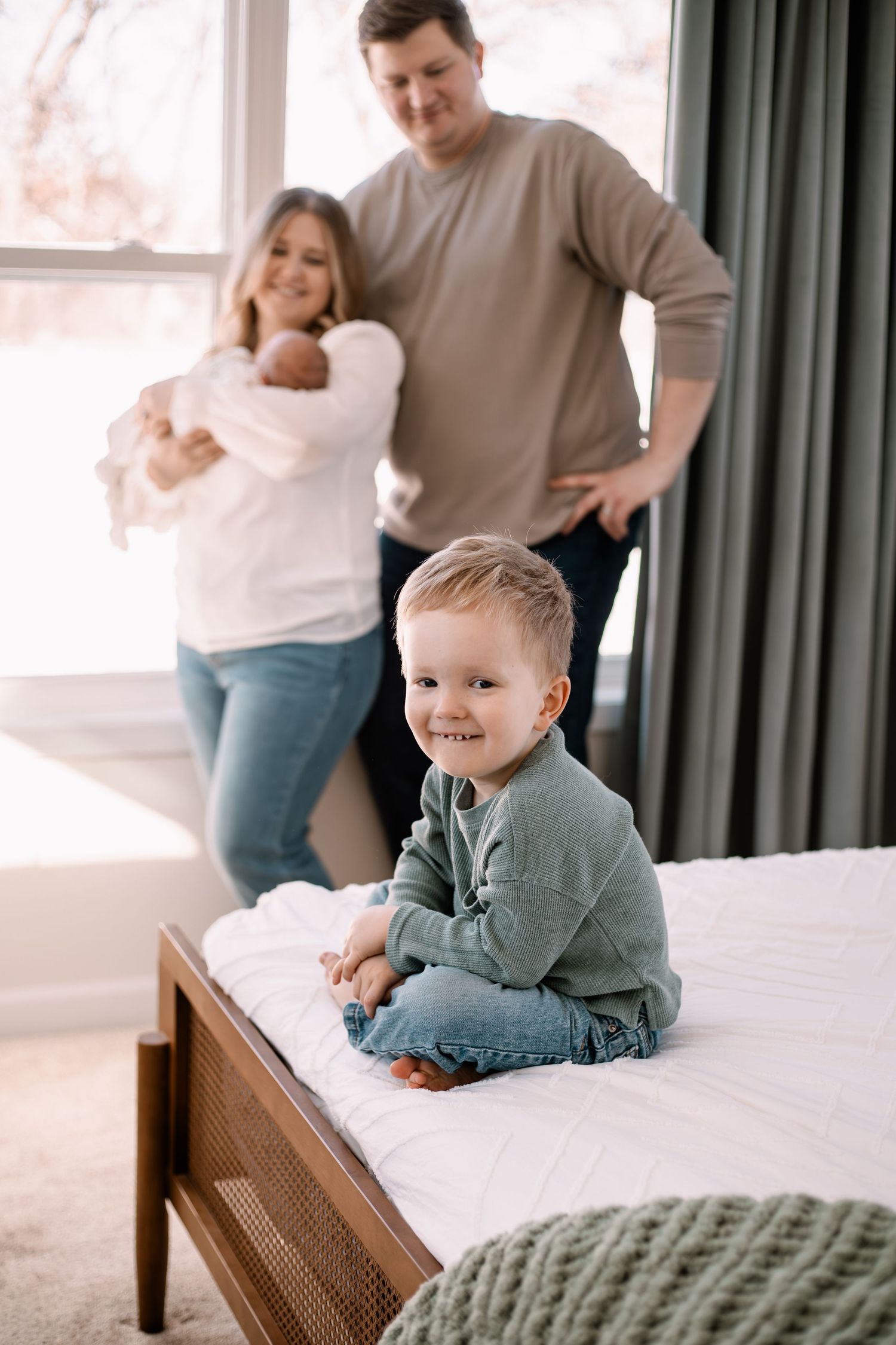 Happy toddler boy sitting on bed smiling at camera while parents hold newborn baby in bright bedroom in Lake Elmo newborn session