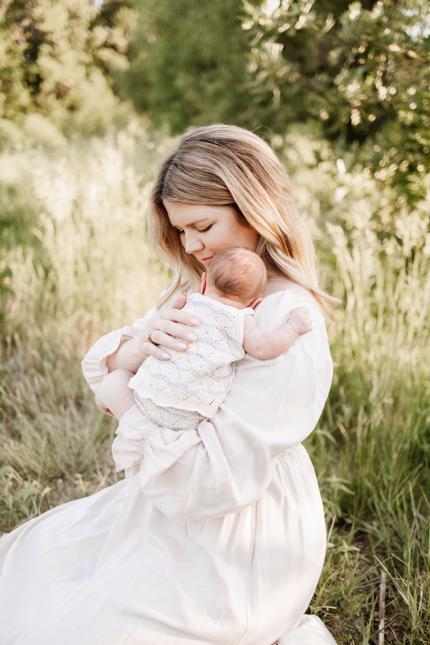 A mother in a white dress tenderly holds her newborn baby outdoors in a sunlit green meadow, taken by a Lakeville newborn photographer.