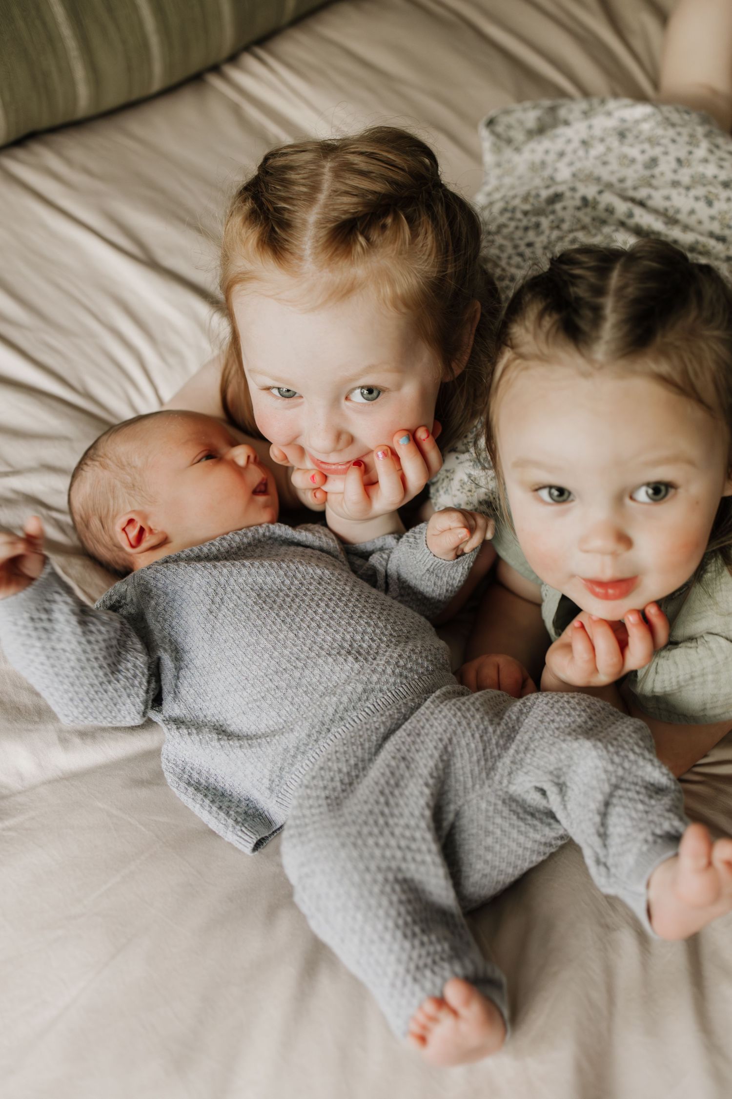 Two toddler girls cuddle with a sleeping newborn baby sibling on a bed, all dressed in soft gray outfits during Minneapolis newborn photos.