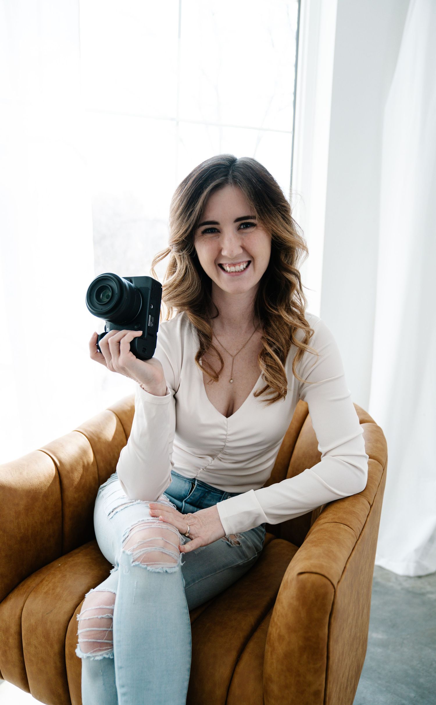 Professional western Wisconsin photographer in white top and ripped jeans sits on brown leather chair holding camera and smiling.