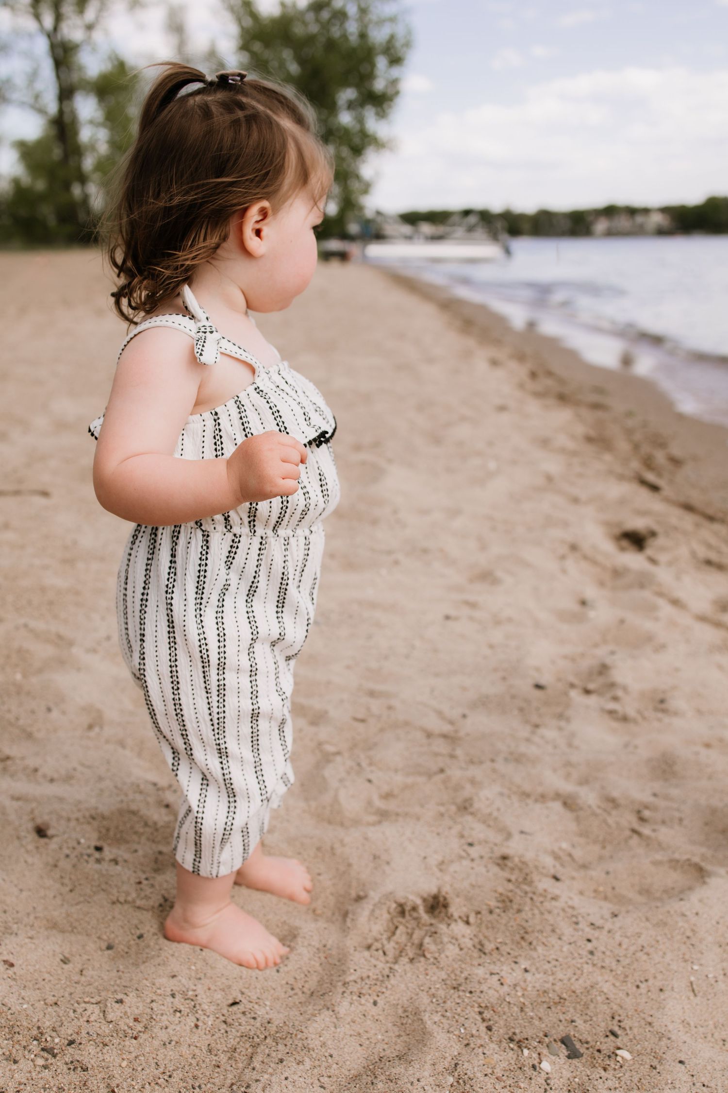 A toddler girl in a striped romper stands barefoot on a sandy beach near the water's edge.
