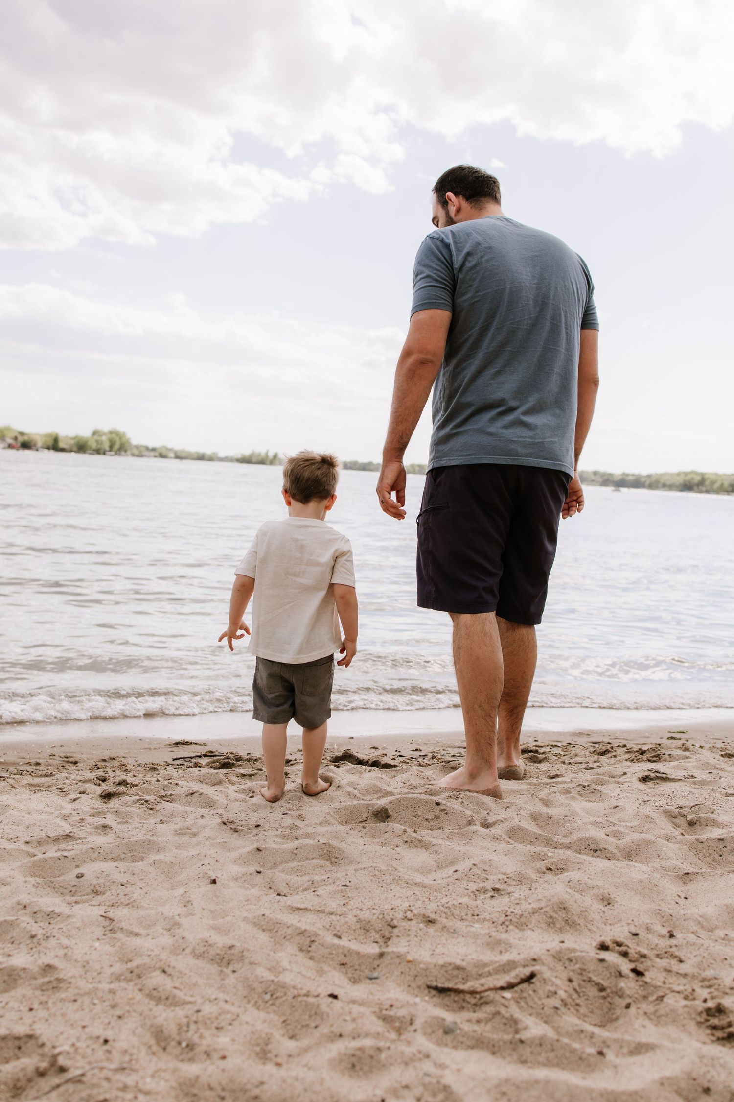 Father and young son standing barefoot on sandy beach shore, viewed from behind, looking out at calm river water.