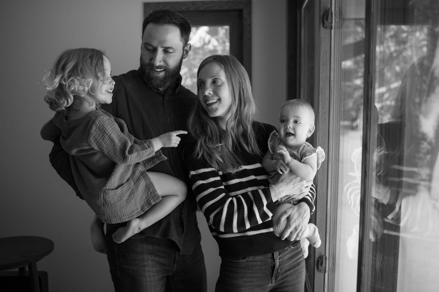 Black and white photo of a happy family with two young children laughing together near a window indoors in Shoreview