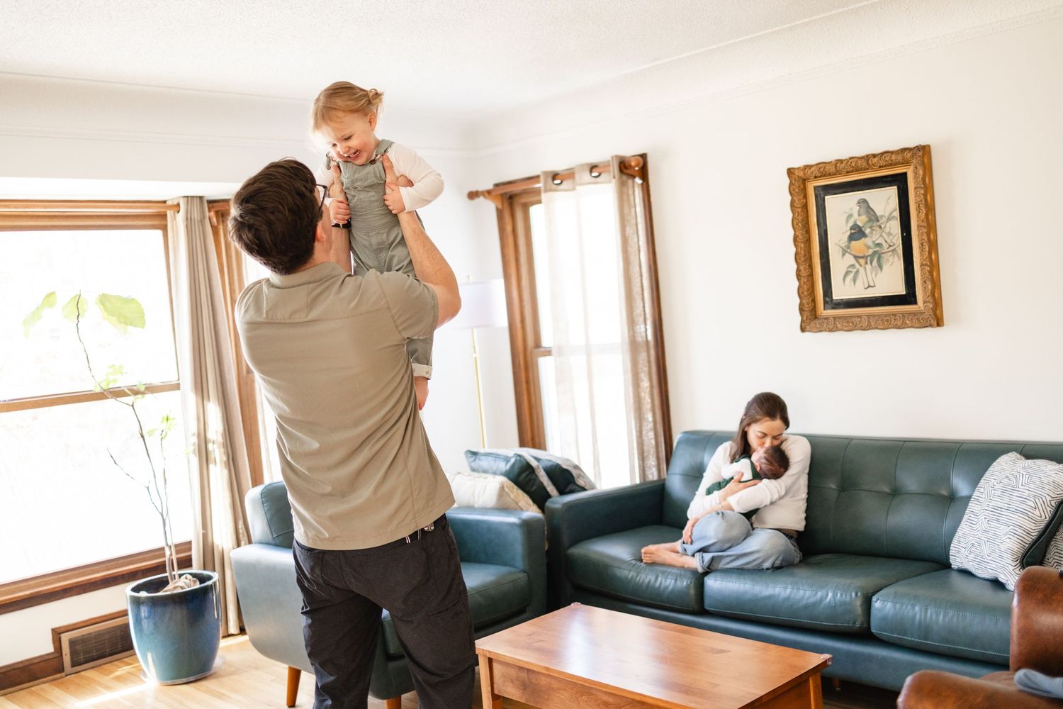 Father lifting baby in living room while mother relaxes on blue sofa in bright family home in Minneapolis