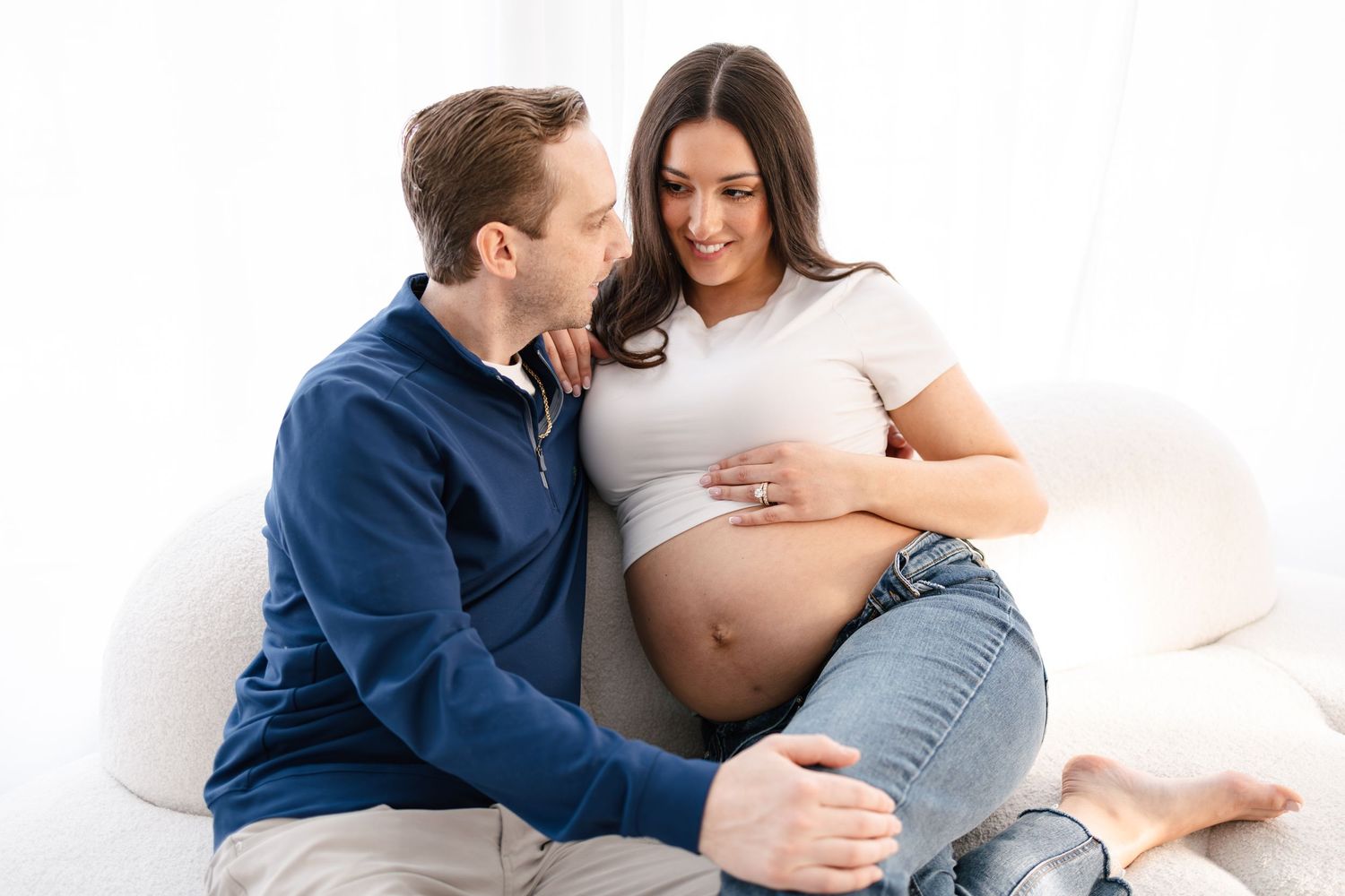 Expecting couple sitting on white couch, man looking lovingly at pregnant woman cradling her belly, photo taken by Woodbury photographer