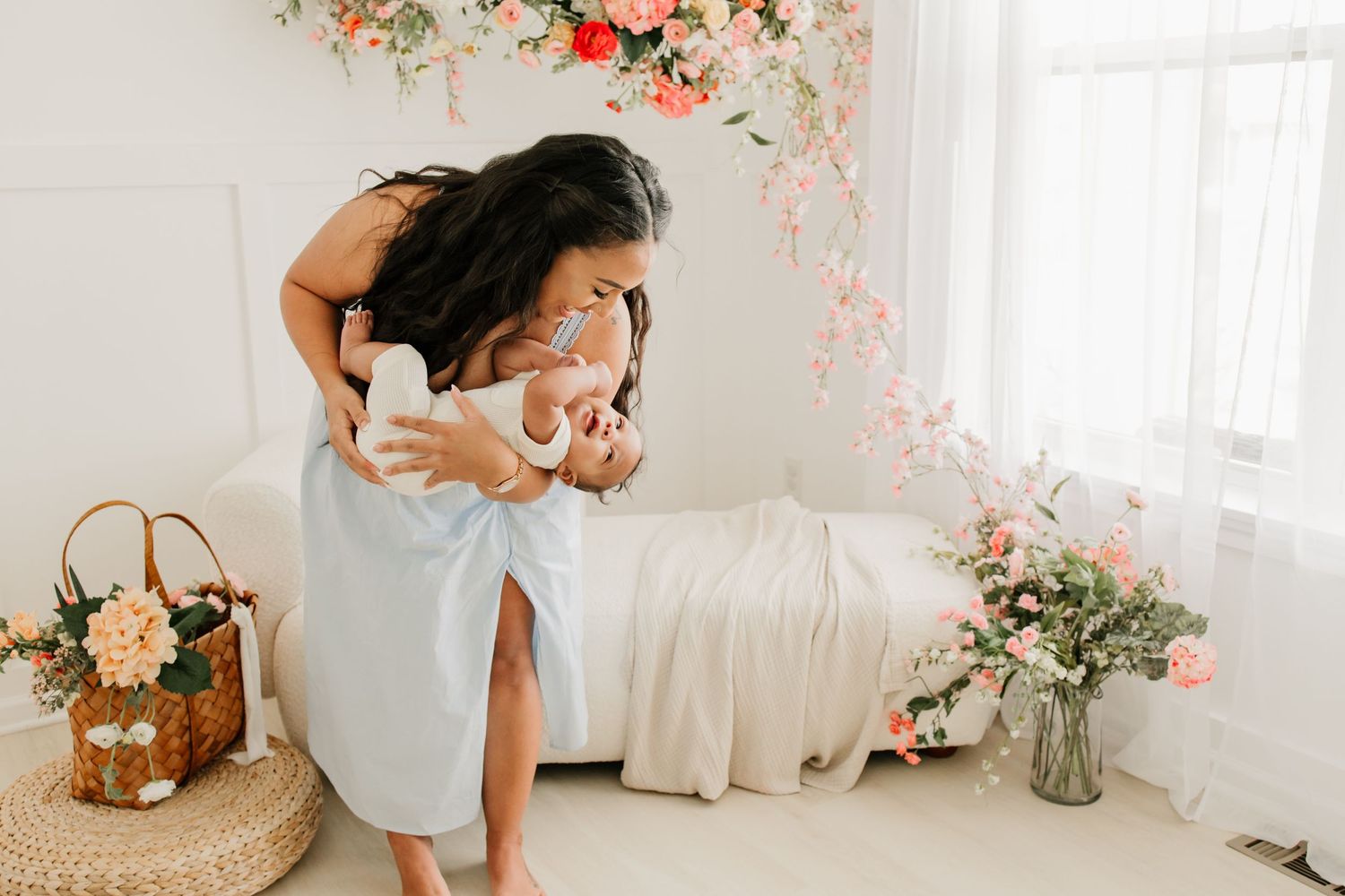 Mother in blue dress lovingly holds newborn baby in bright floral studio with pink blooms and soft natural light in Lakeville