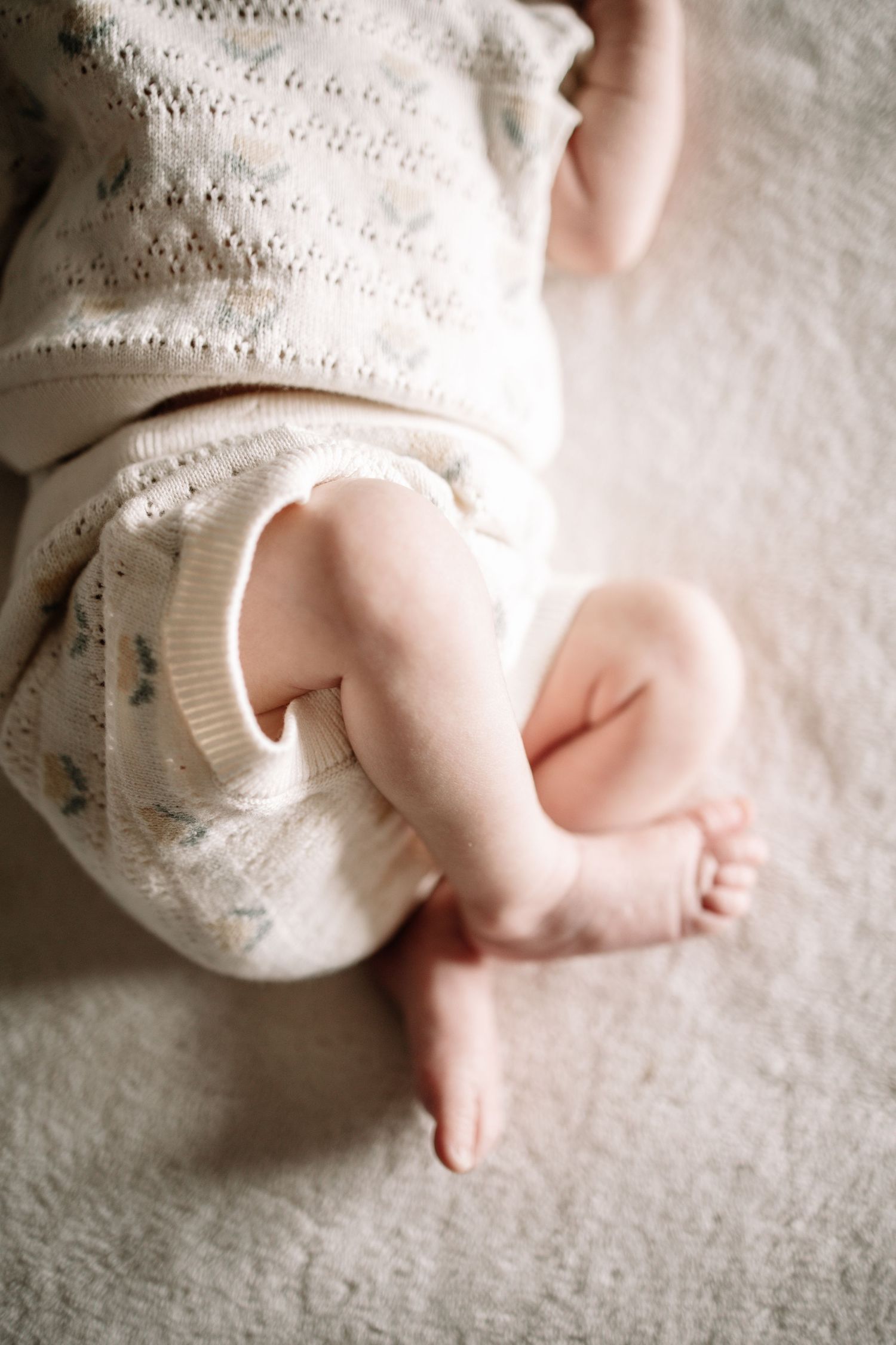 Close-up of a newborn baby's tiny feet and legs in a soft white knit outfit, lying on a cream blanket.