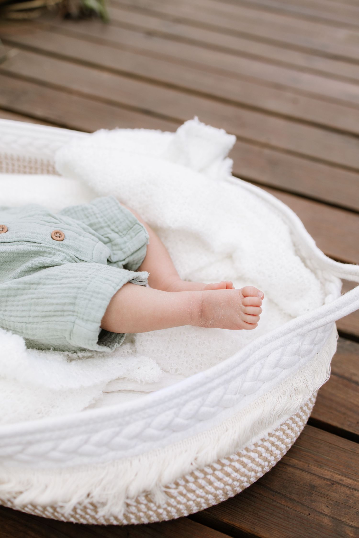 Baby resting in a white woven basket with soft blanket, wearing sage green outfit, tiny bare feet visible on wooden deck.