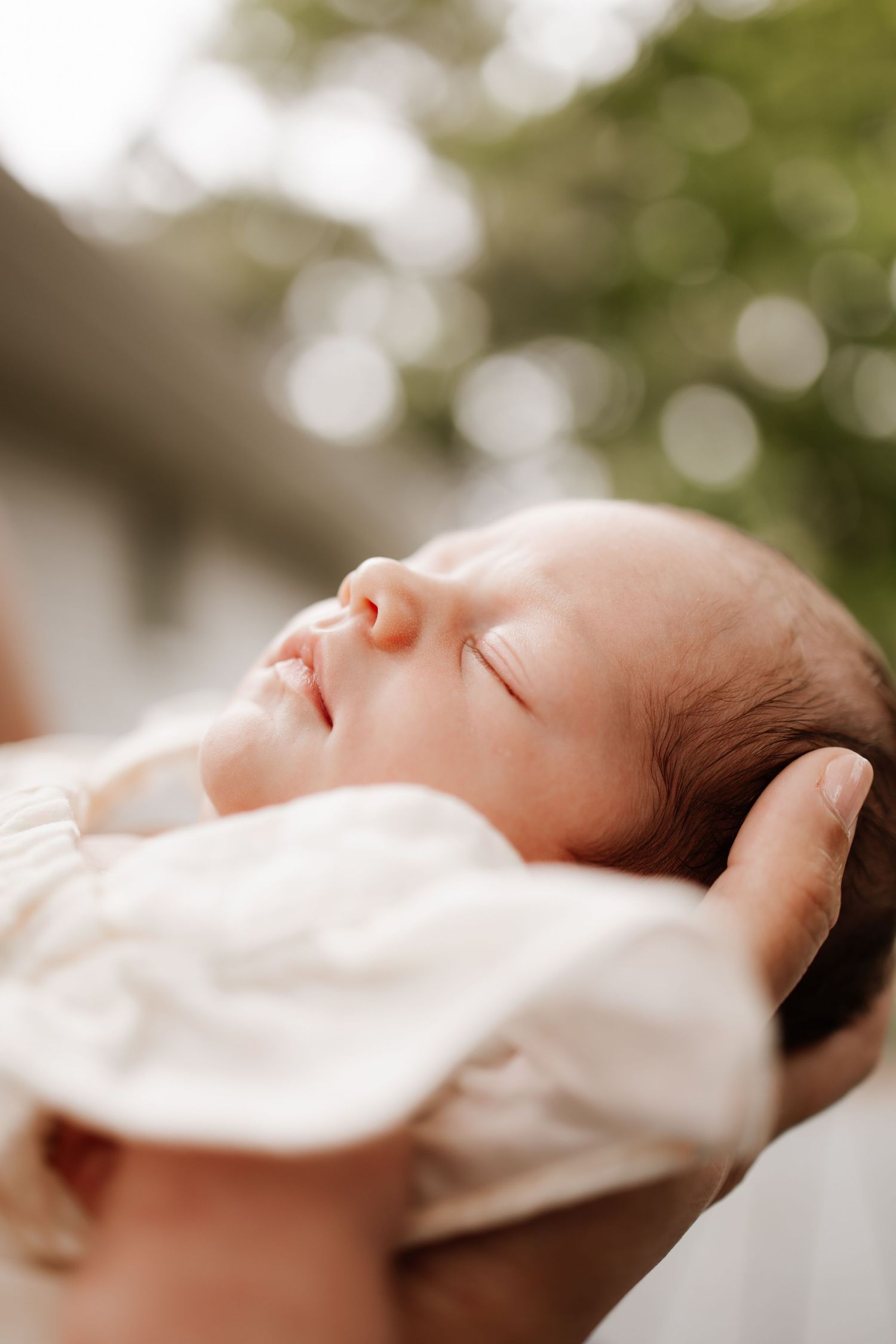 A peaceful newborn baby sleeping in a parent's gentle hands outdoors at in-home newborn session in Woodbury, dressed in a soft white outfit.