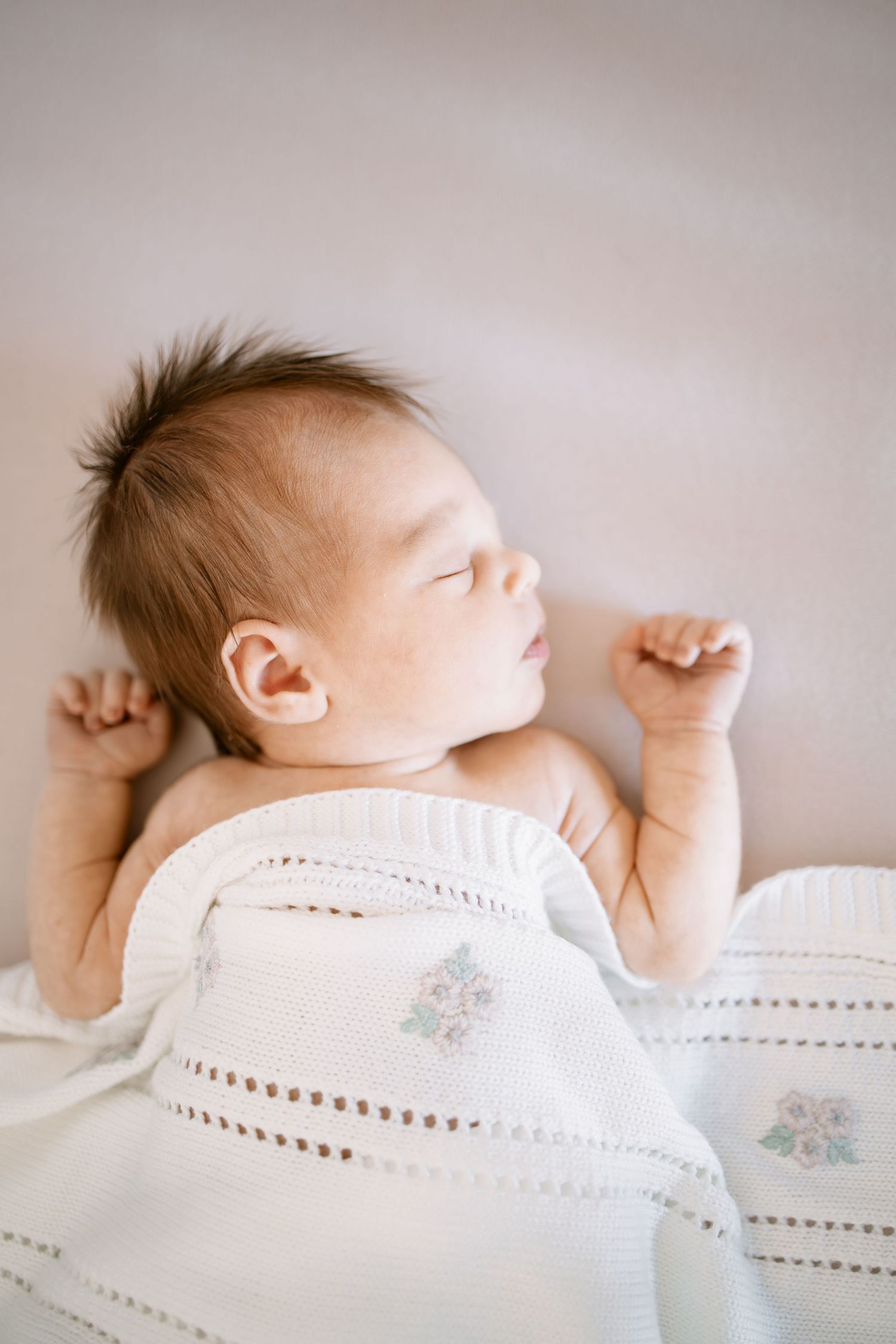 Peaceful newborn baby sleeping under a white floral blanket with tiny fists curled during in-home newborn session by Woodbury photographer.