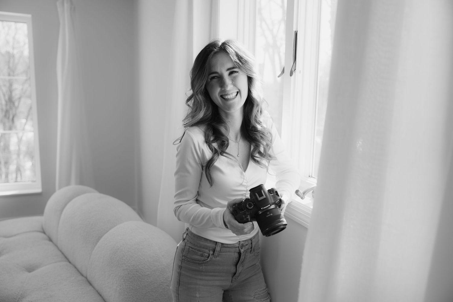 Black and white photo of a Woodbury photographer holding a camera while standing by a window in a bright interior room.
