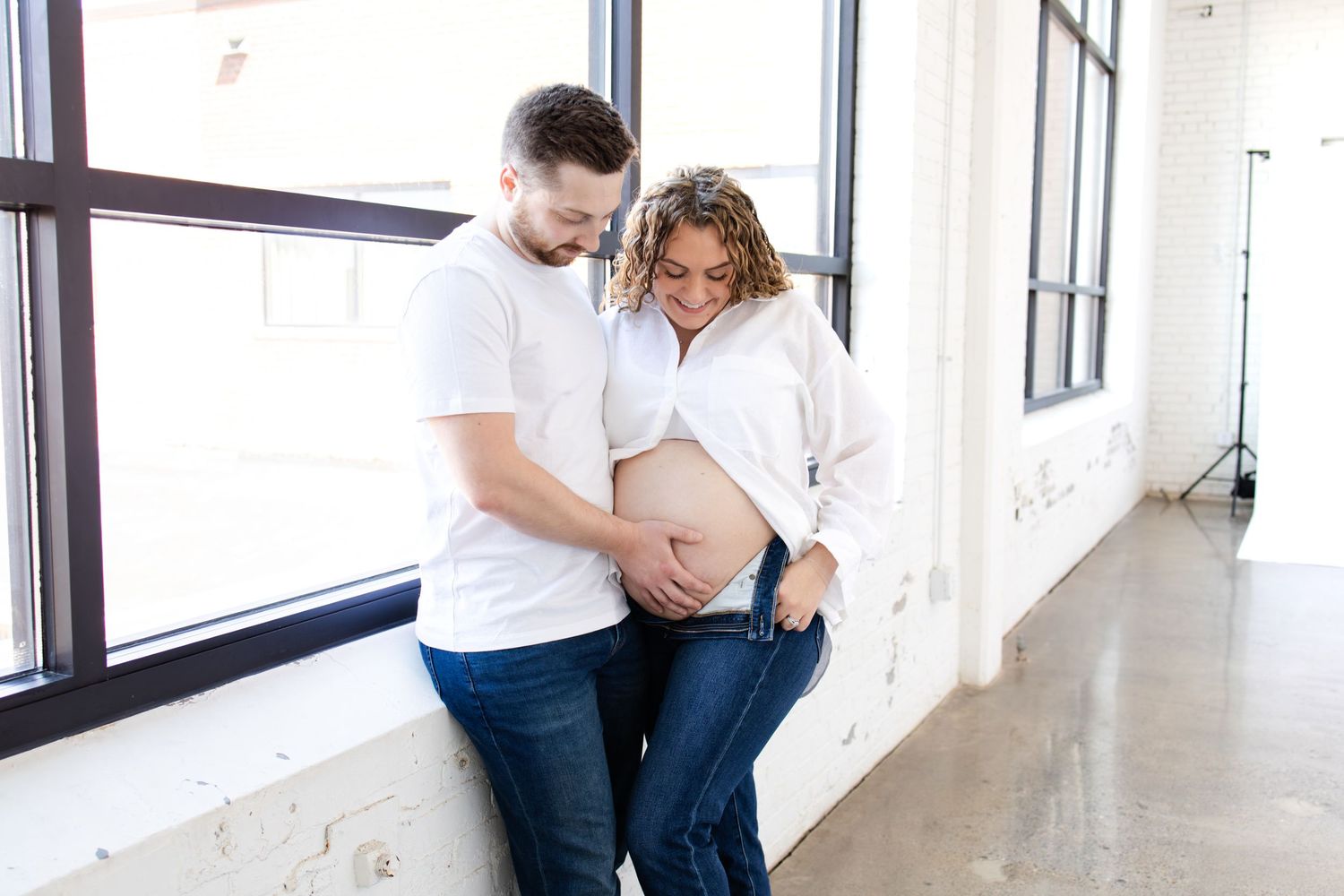 Expecting couple in casual attire tenderly looking down at pregnant belly near bright windows during Minneapolis photo session