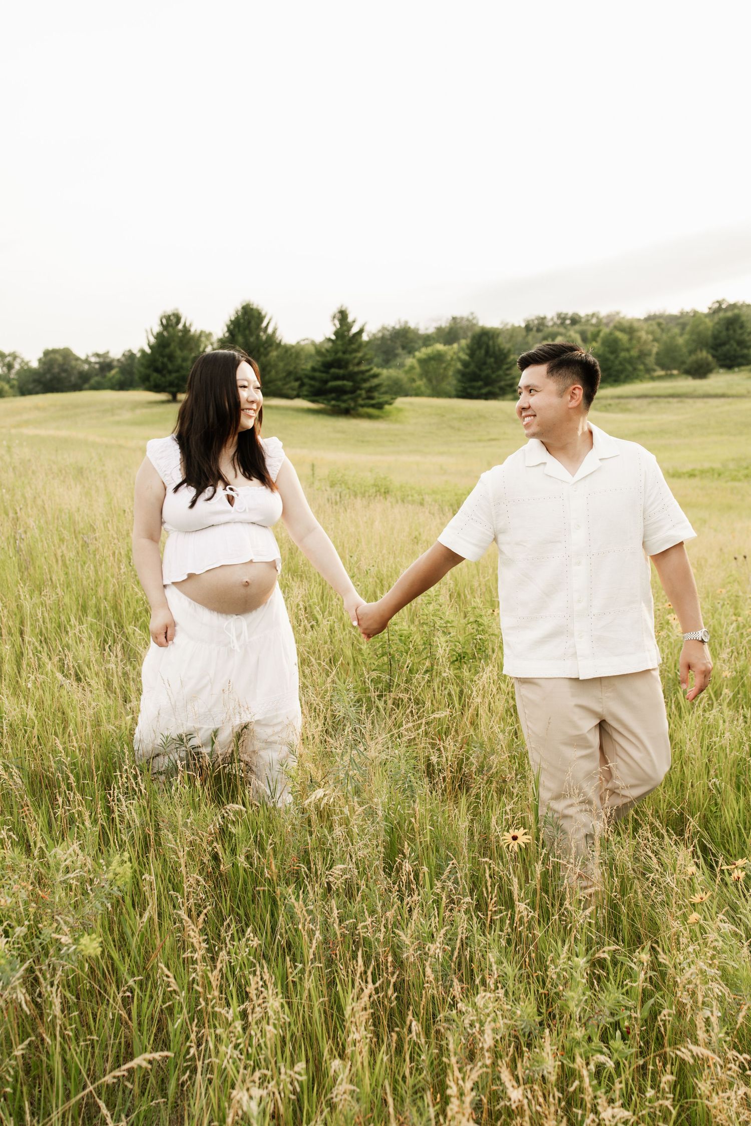 Pregnant couple holding hands walking through a lush green field, wearing white and beige outfits.