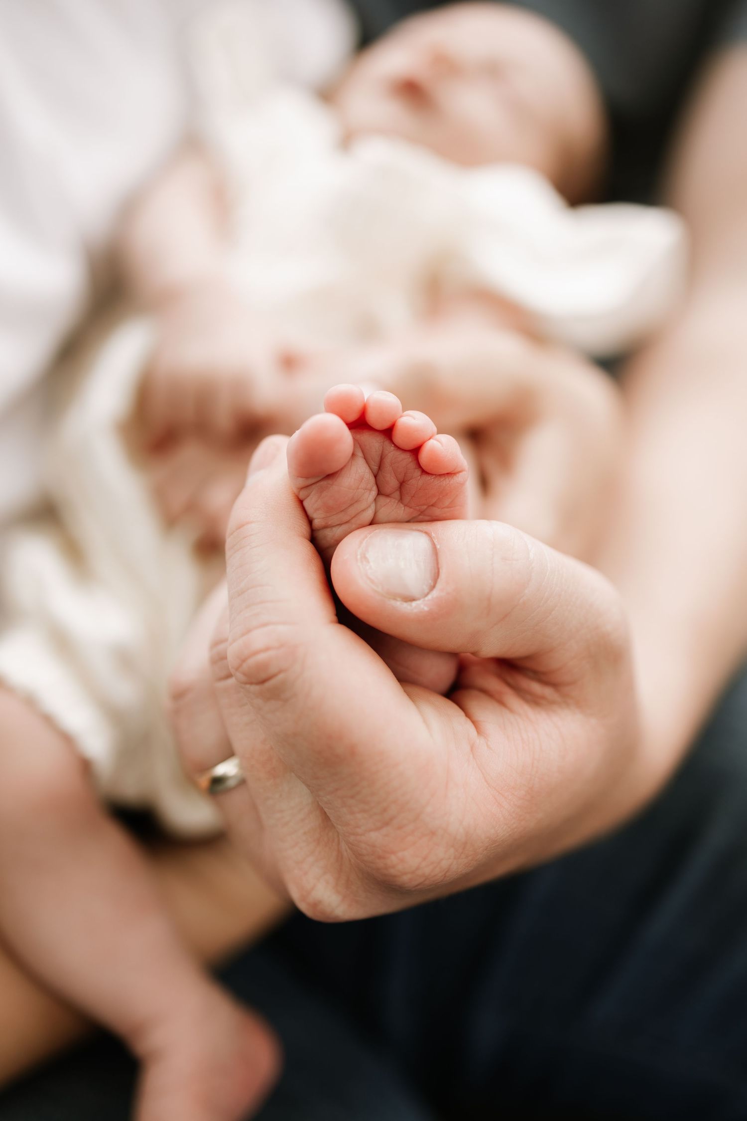 Close-up of an adult hand gently holding tiny newborn baby feet, with infant resting in background.