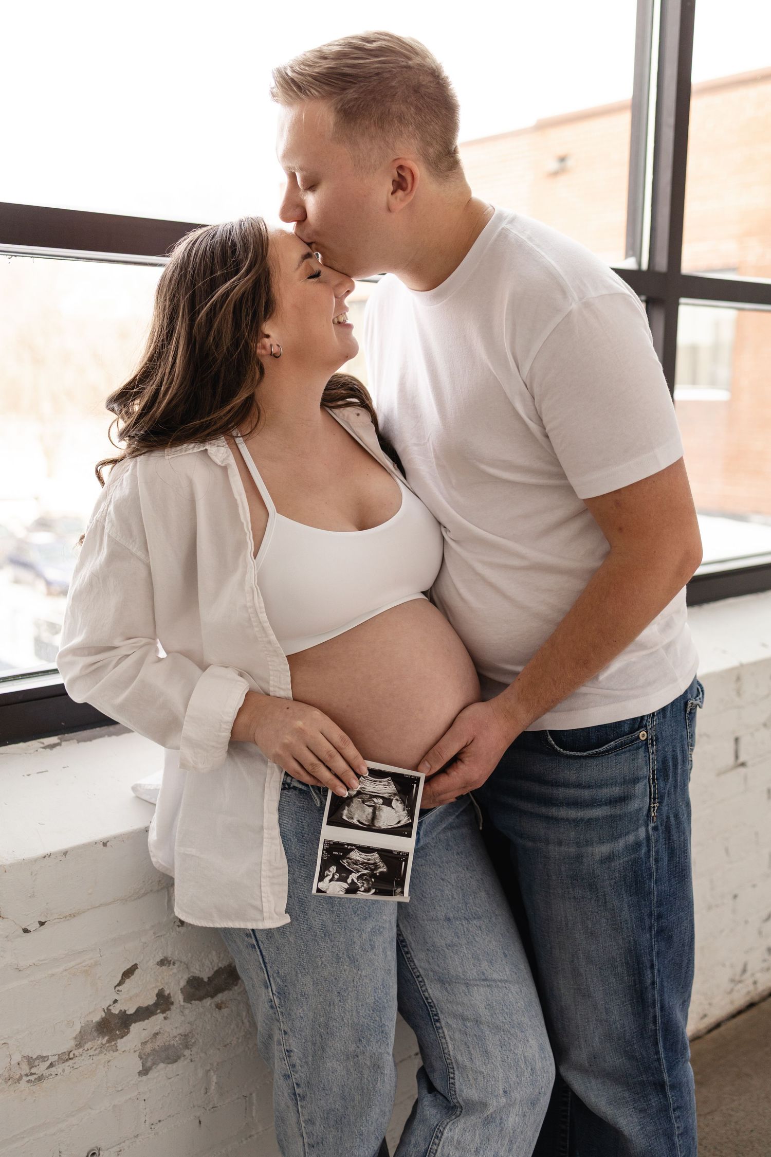 Couple sharing a kiss while pregnant woman holds ultrasound photo, standing by window in casual white tops and jeans in Minneapolis