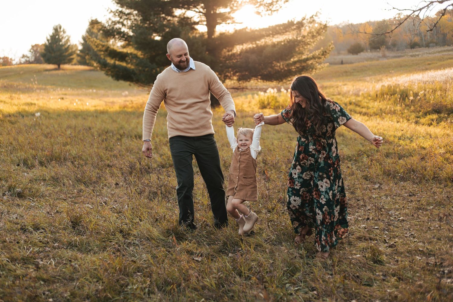 Family of three walking in golden hour field, parents swinging toddler between them outdoors during fall mini session in Lakeville, MN.
