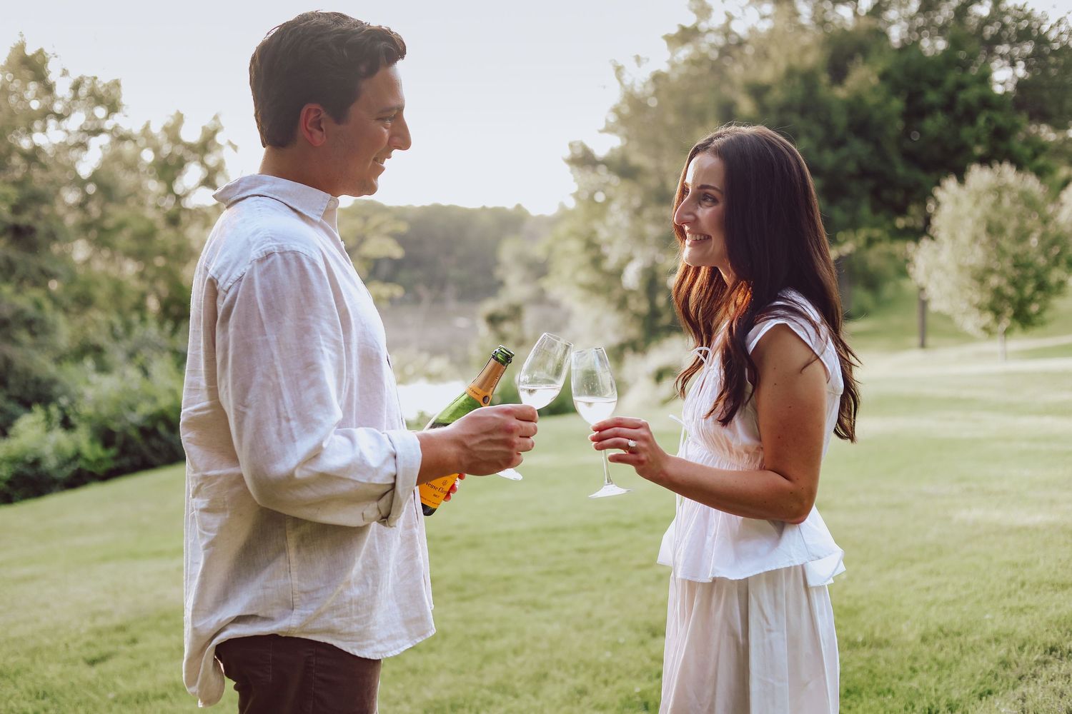 A couple in white clothes shares a romantic moment while holding drinks in a sunlit garden setting taken by Woodbury engagement photographer