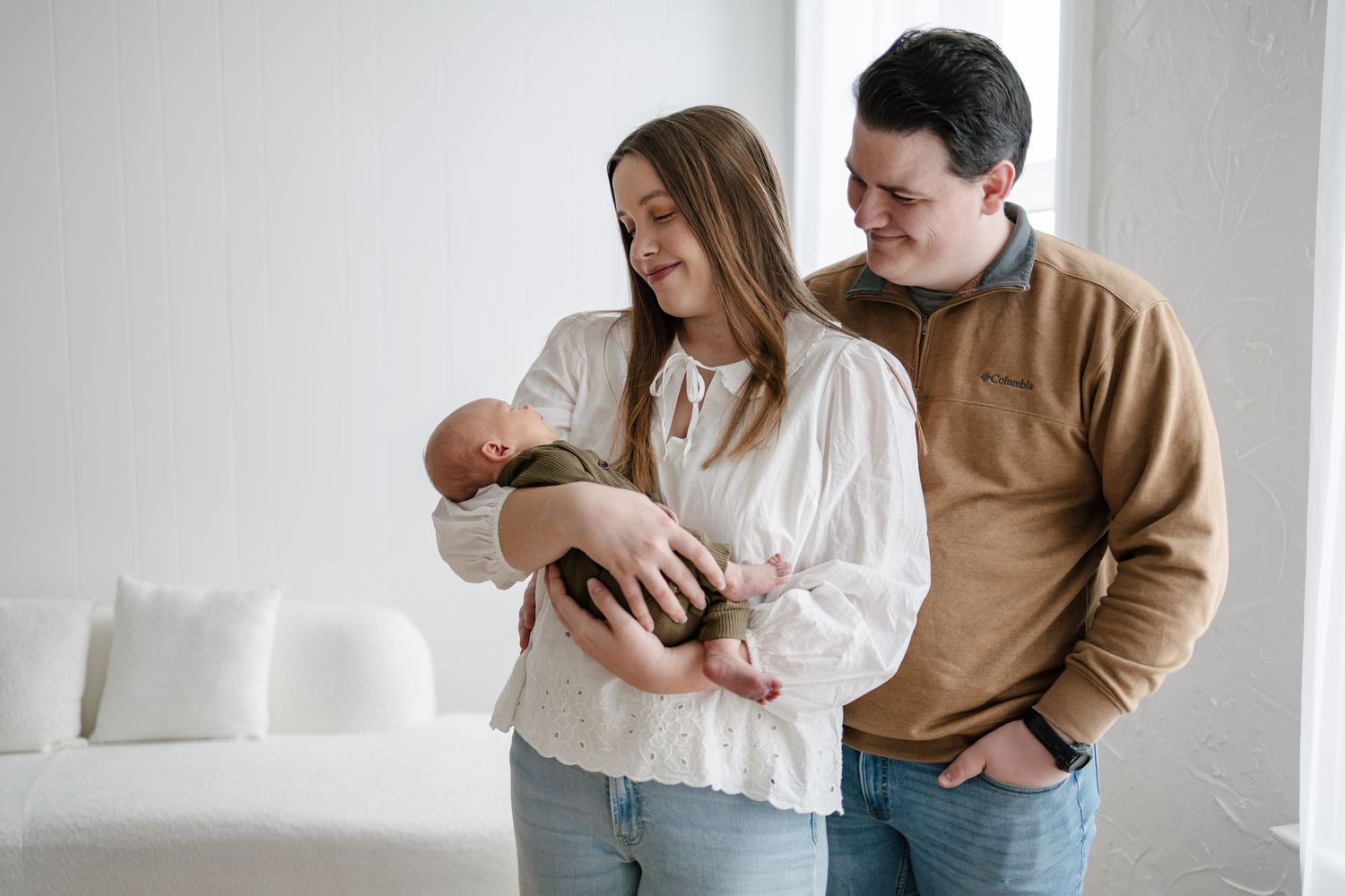 A couple smiles while cradling their newborn baby in a bright white room during Woodbury studio newborn session