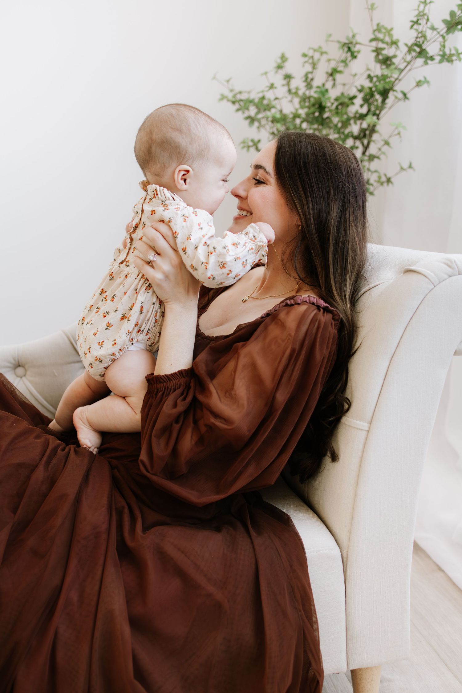 A mother in a brown dress holds her baby close, smiling tenderly while seated in a white chair taken by Woodbury motherhood photographer.