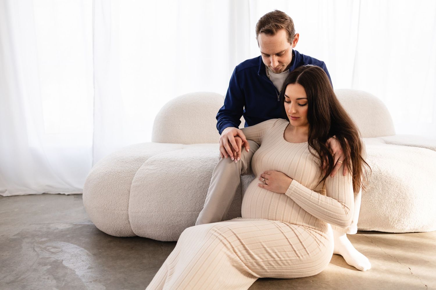 A pregnant couple casually sitting against a white couch, looking down at a pregnant belly in a natural light studio setting in Twin Cities.