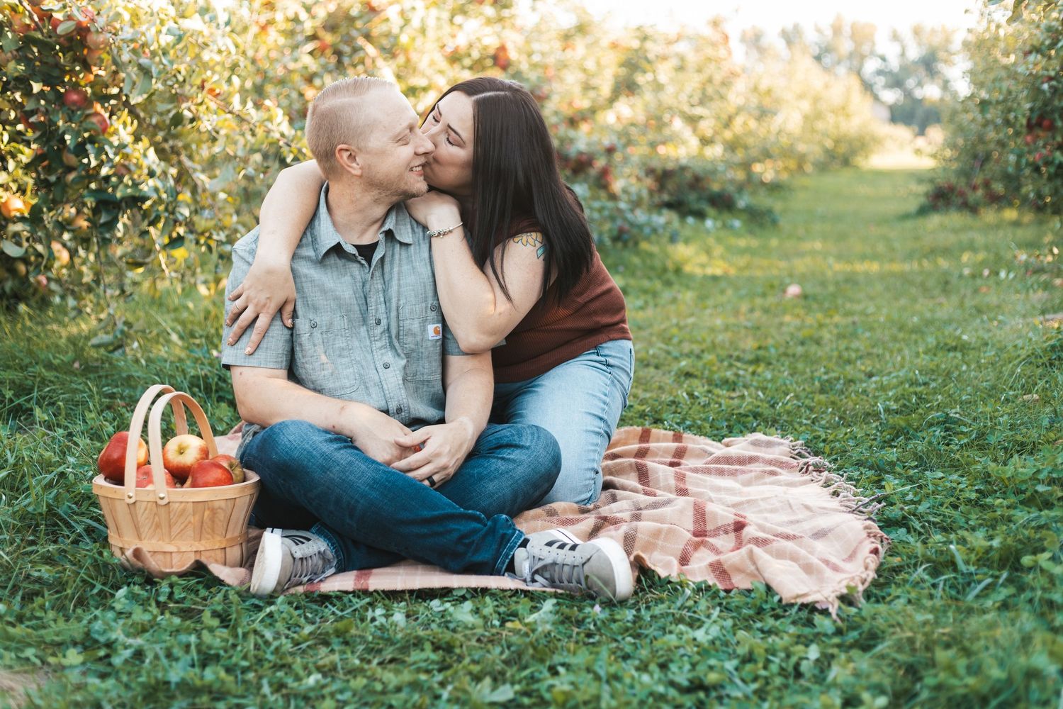 A romantic picnic scene between apple tree rows, with a woven basket and blanket spread on green grass during Lakeville orchard mini session