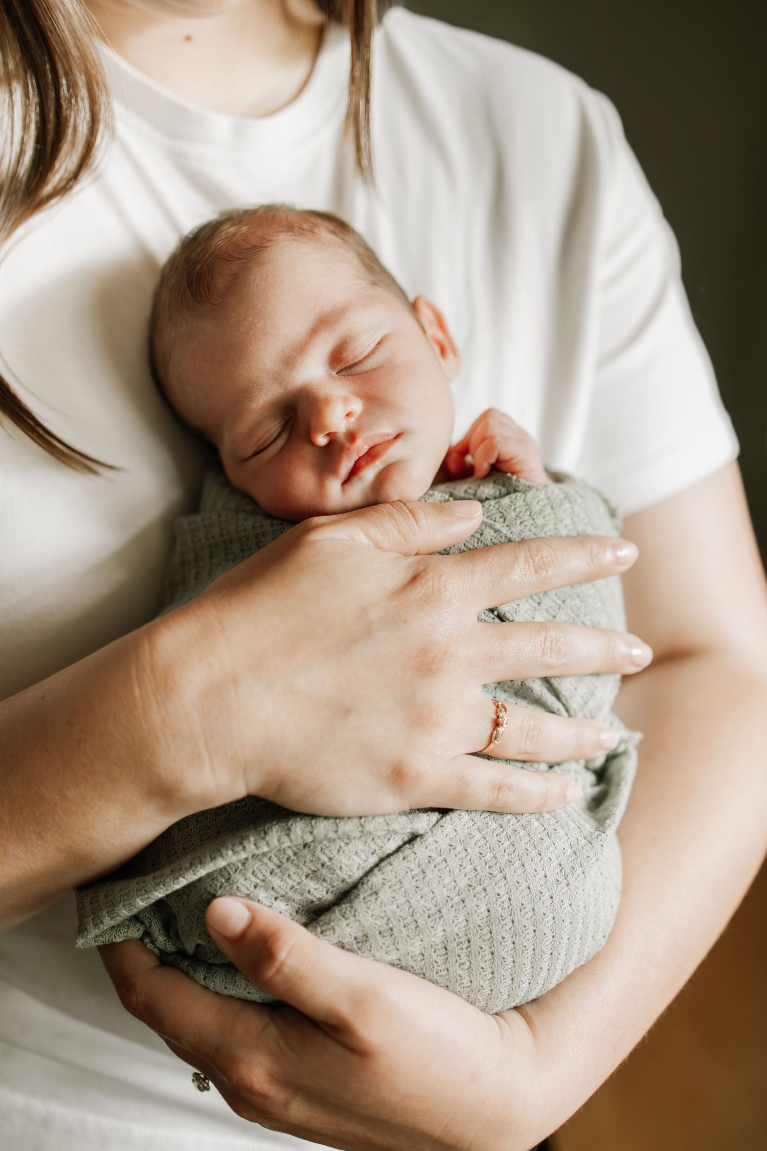 A newborn baby swaddled in a sage green wrap, sleeping peacefully in a mother's gentle arms during Twin Cities in home newborn photos.