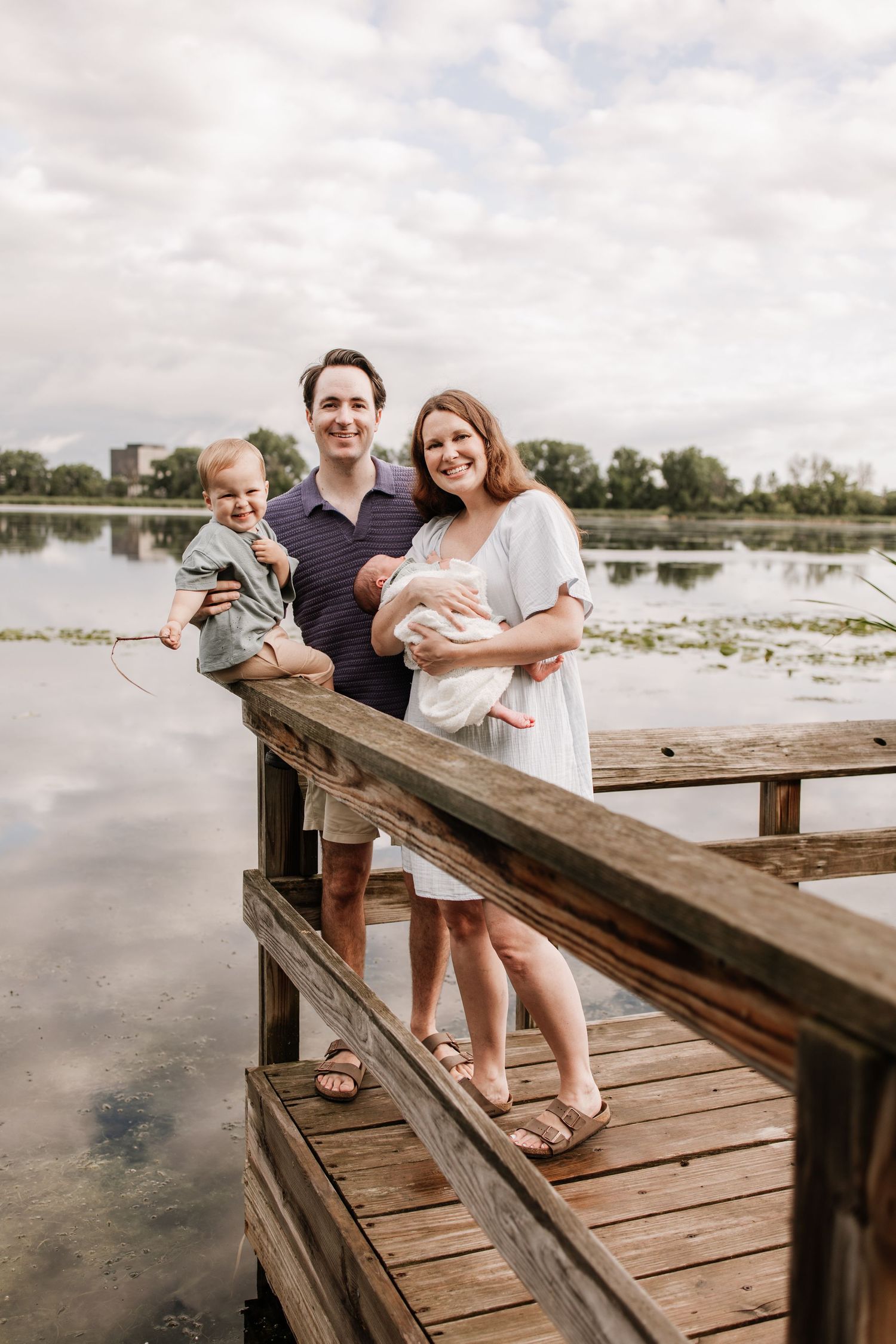 A family of four poses on a wooden dock by a calm lake, with parents holding their newborn baby in an outdoor newborn session in Minneapolis
