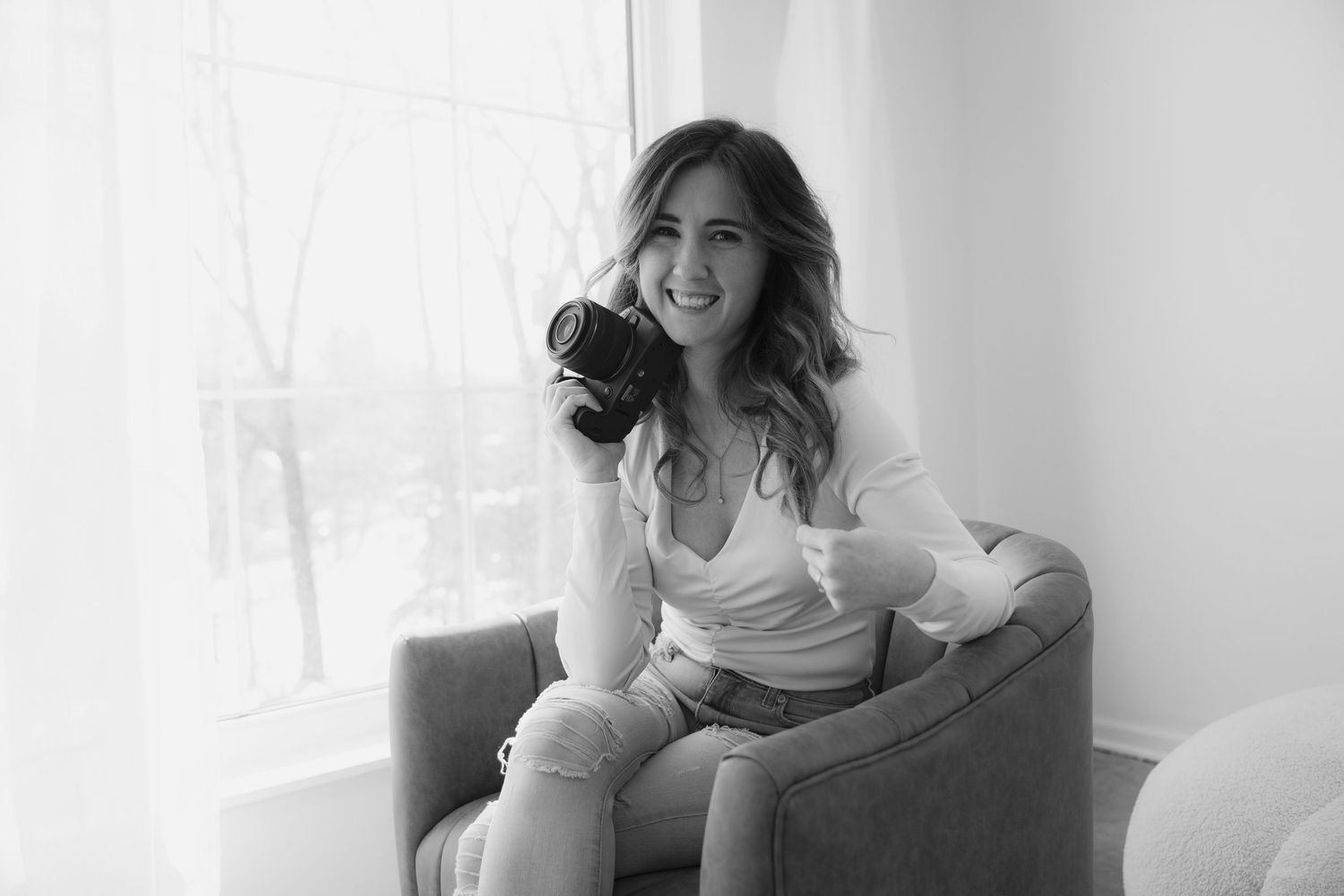 A joyful Woodbury photographer sits on a chair in a bright room while wearing a white top and casual attire.