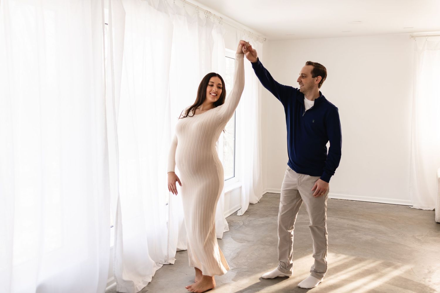 A pregnant couple enjoys a sweet twirl as they dance in a bright natural light studio setting, taken by Twin Cities maternity photographer.