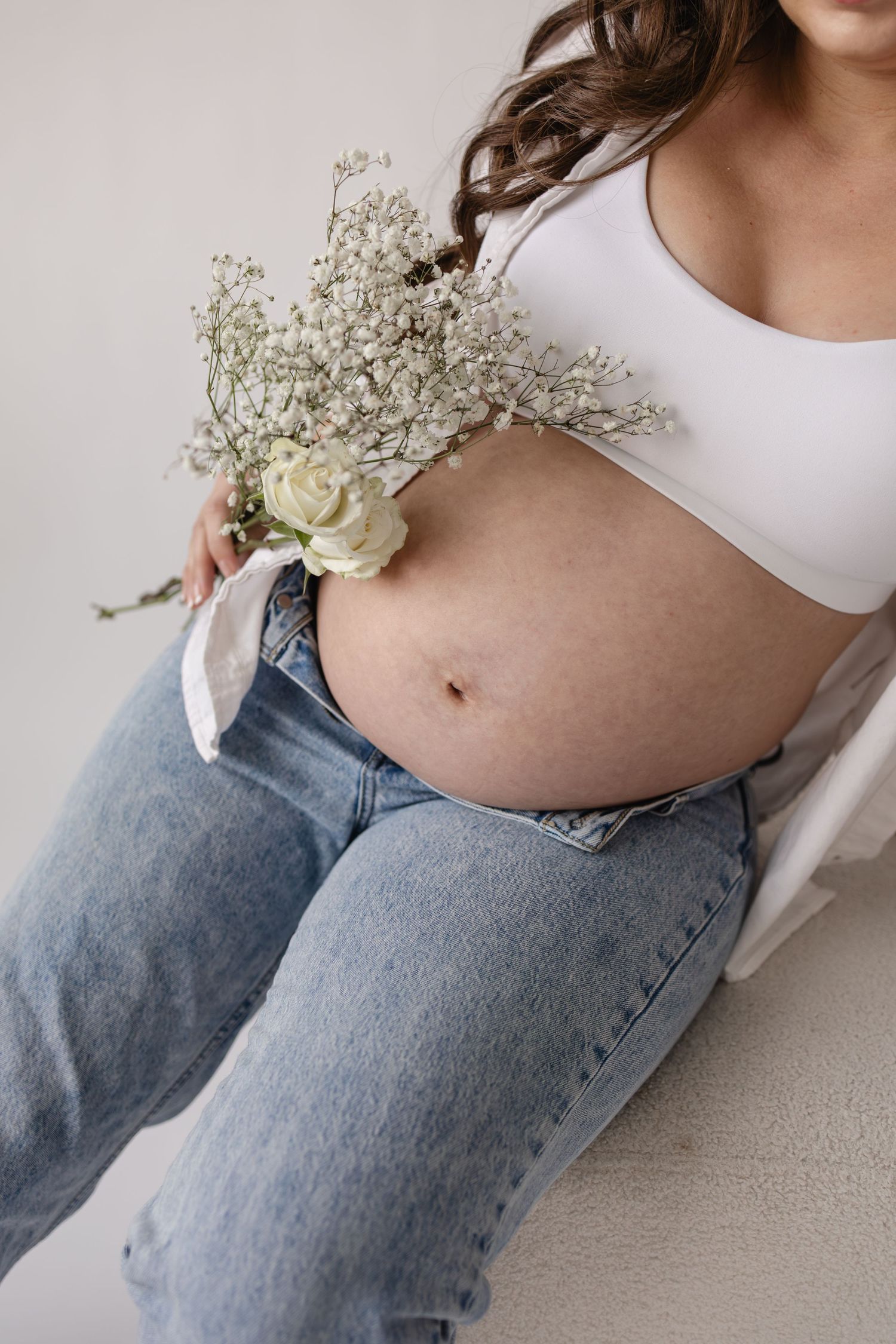 Pregnant woman in white top and jeans holding delicate white flowers and roses against her bare baby bump in Minneapolis maternity session