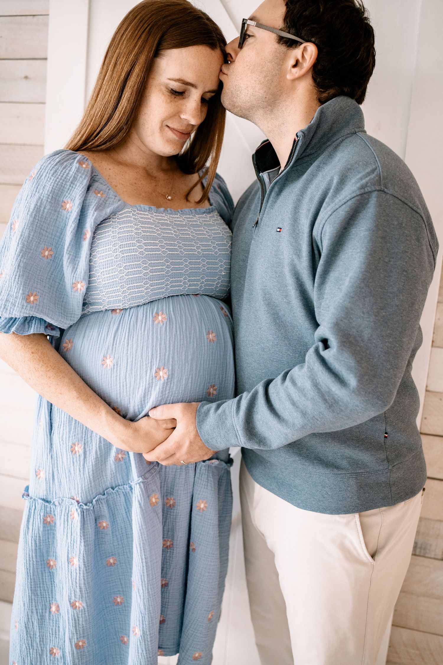 Woodbury family photographer captures photo o man kissing pregnant woman's temple as they hold her baby bump together, both smiling warmly.