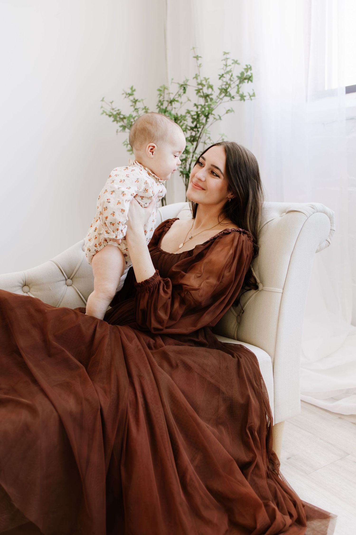 A mother in a brown dress sits in a white chair, lovingly holding her baby in a bright, airy room during motherhood photo session.