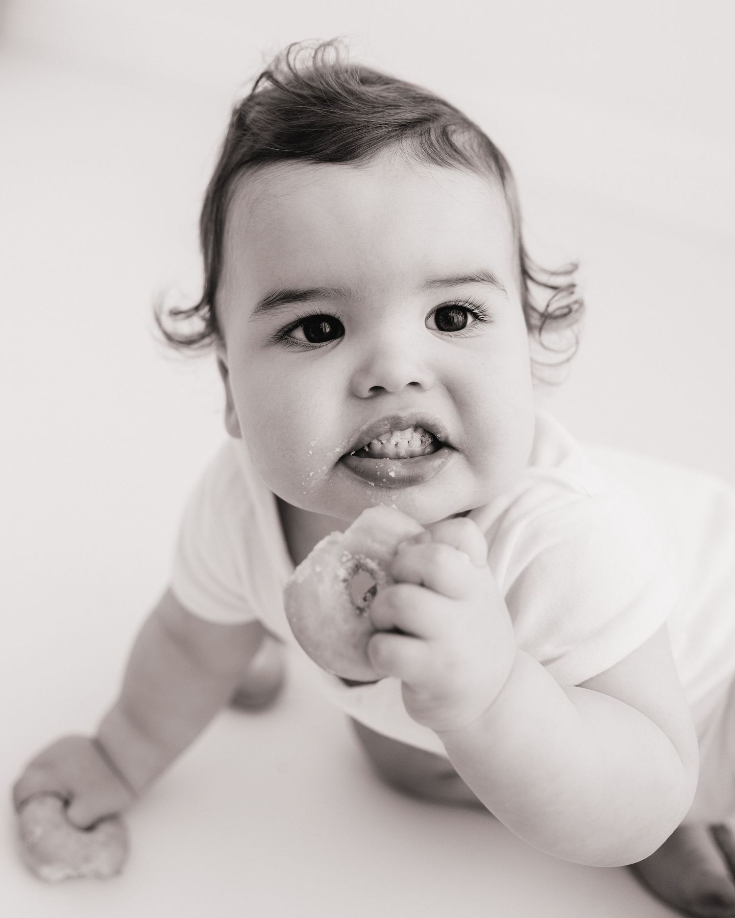 wide eyed 1 year old holding a donut in each hand looks into the camera while crawling around Ayrshire studio during milestone session