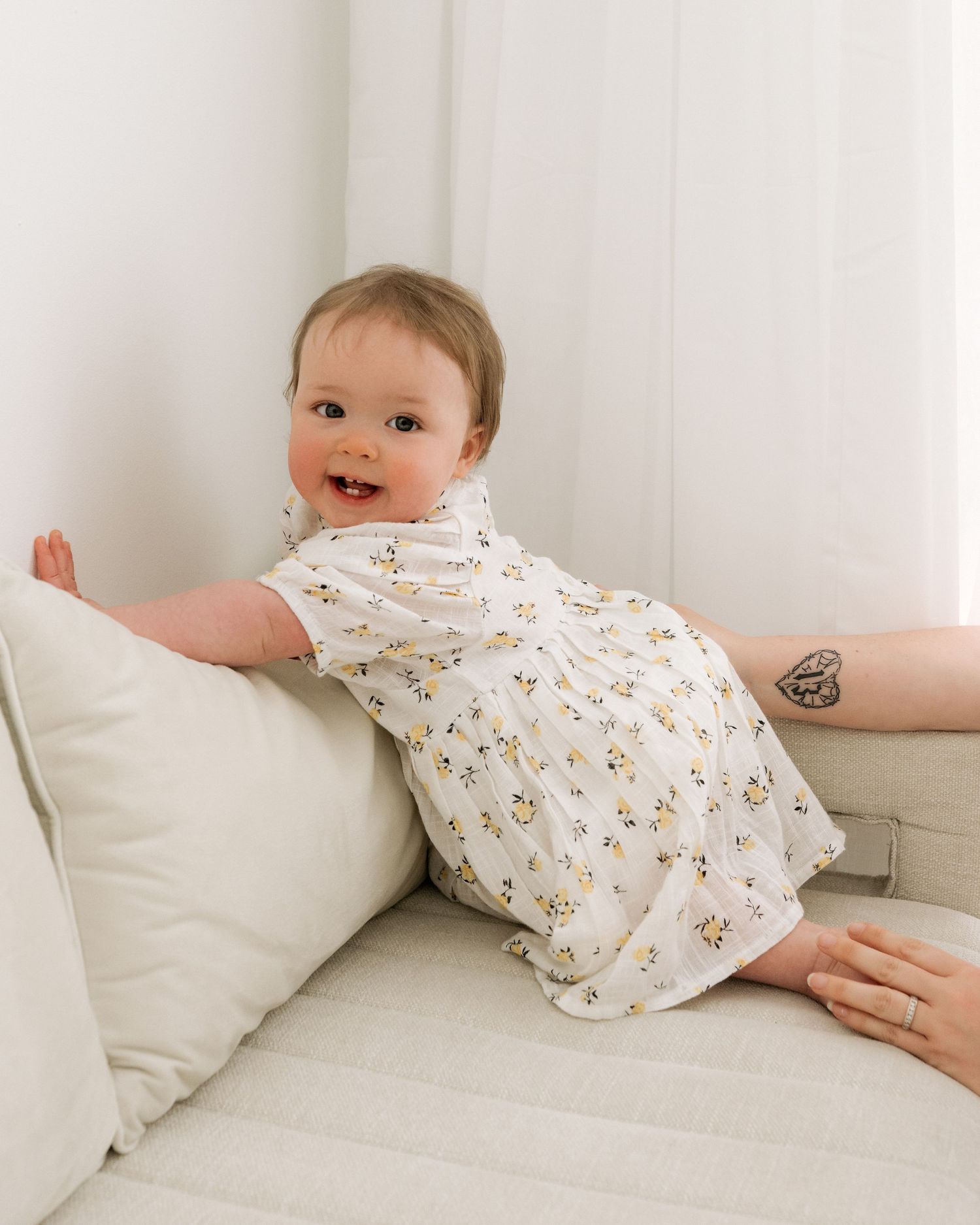 kneeling on sofa, reaching for the wall, baby smiles into camera as mum watches on during 1st birthday photoshoot in Ayrshire