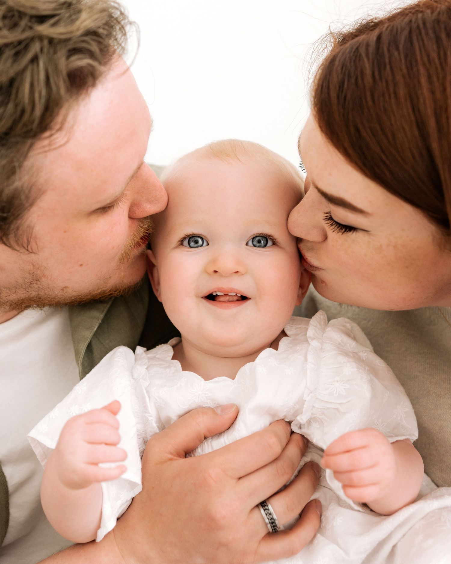 first time parents kiss their almost 1 year old on the cheek as she looks directly into the camera