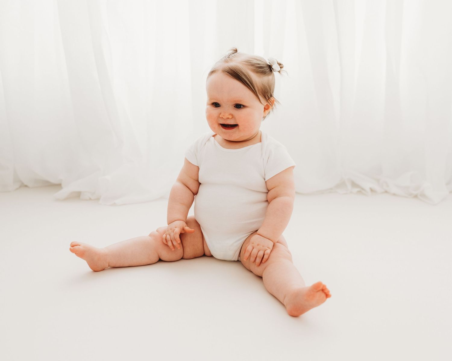 smiling baby sits in white vest on floor of all-white photo studio in Ayrshire during her 1st birthday photoshoot with Faye McRae