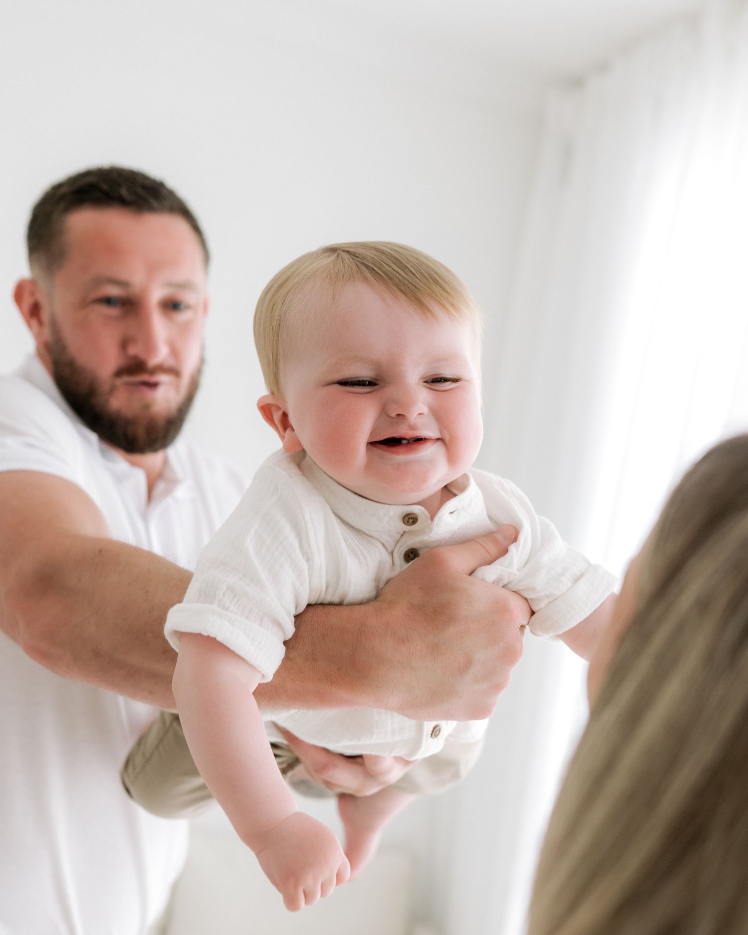 dad swooshes his 1 year old son to his mum, baby is smiling and laughing during baby-led milestone session in Faye McRae's Ayr studio