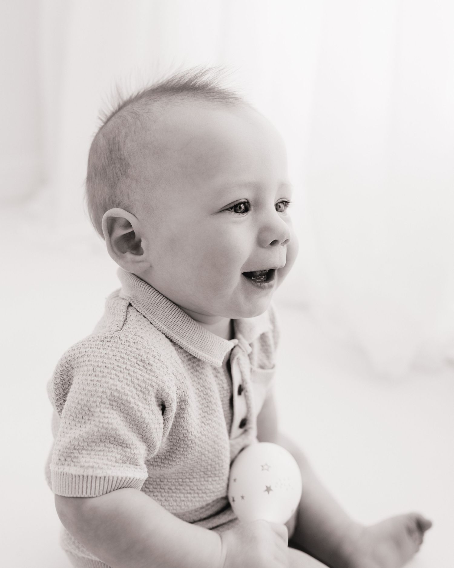 smiling 9 month old baby holds a maraca and his wide eyes looking at his mum, taken by Ayrshire baby photographer Faye McRae
