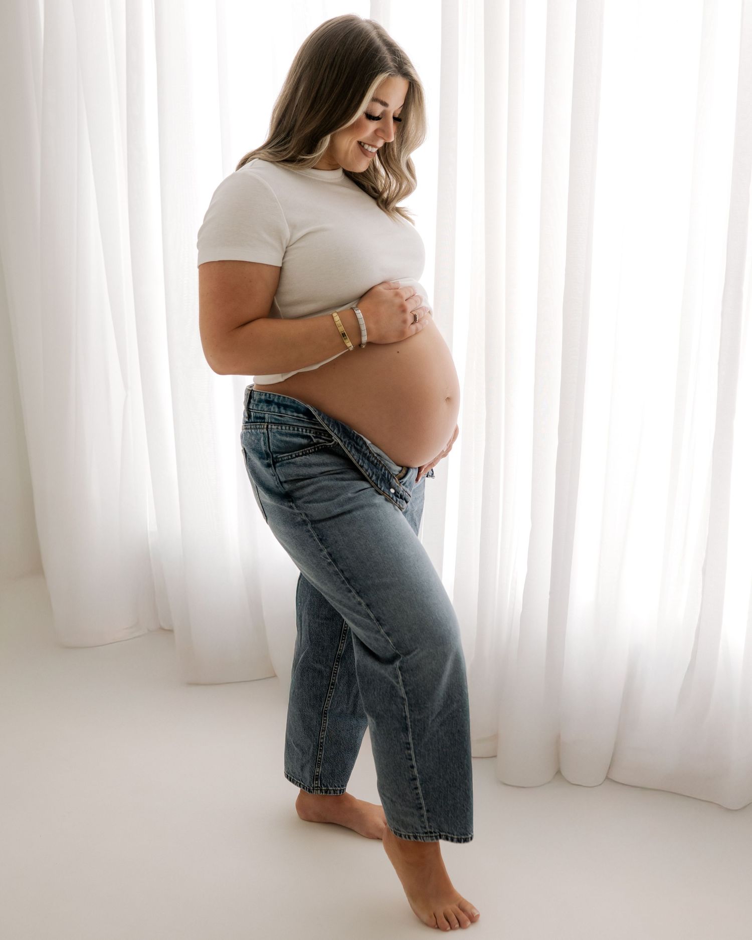 wearing simple blue jeans and white t-shirt, smiling first time mum looks down at her bump that she is cradling with her hands in Ayrshire