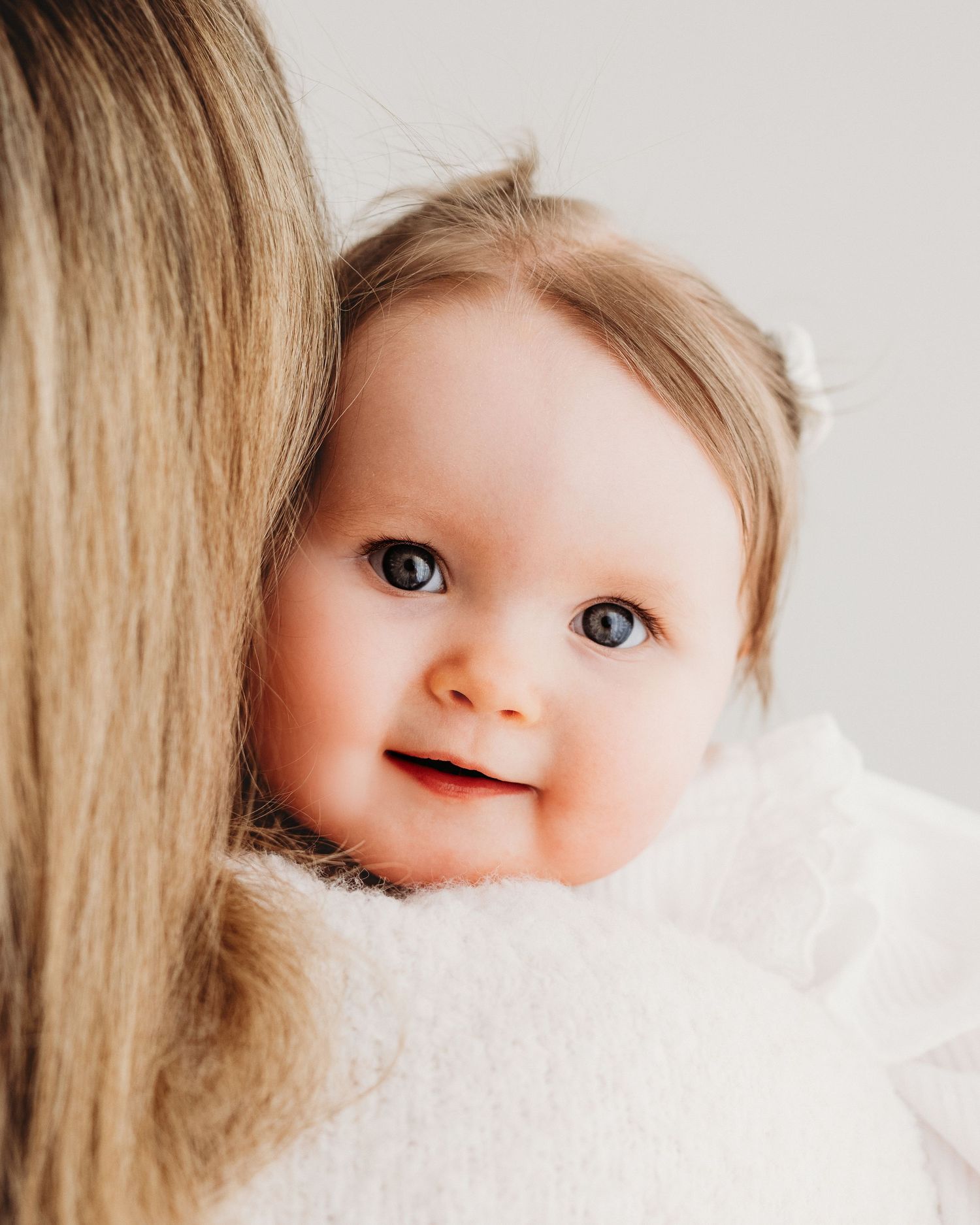 wide eyed 1 year old girl gazes into the camera over her mums shoulder at her 1st birthday photoshoot in Faye McRae's Ayrshire studio