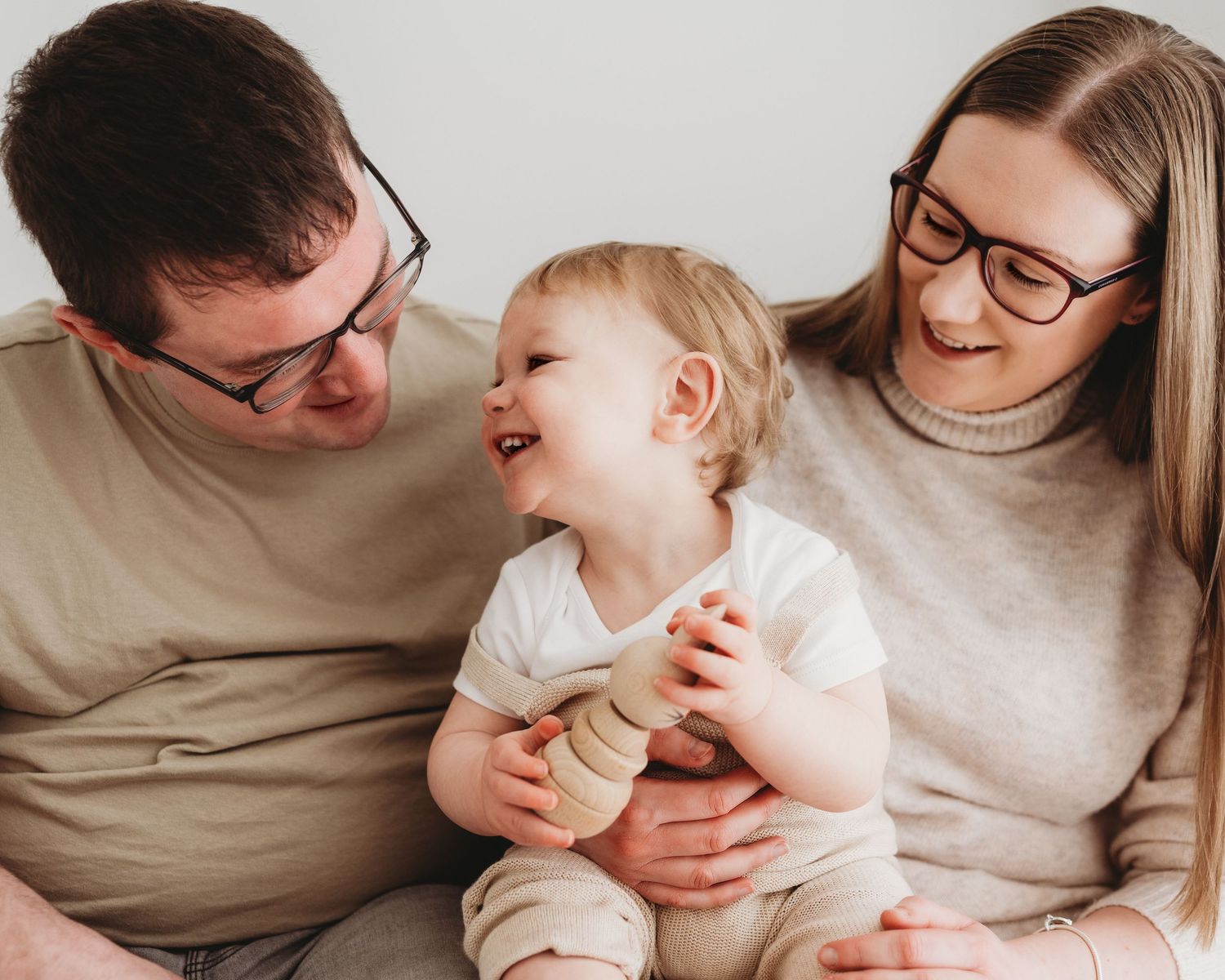 family sit together smiling and laughing, baby holds a wooden toy smiling up at his dad during his 1st birthday photoshoot in Ayrshire