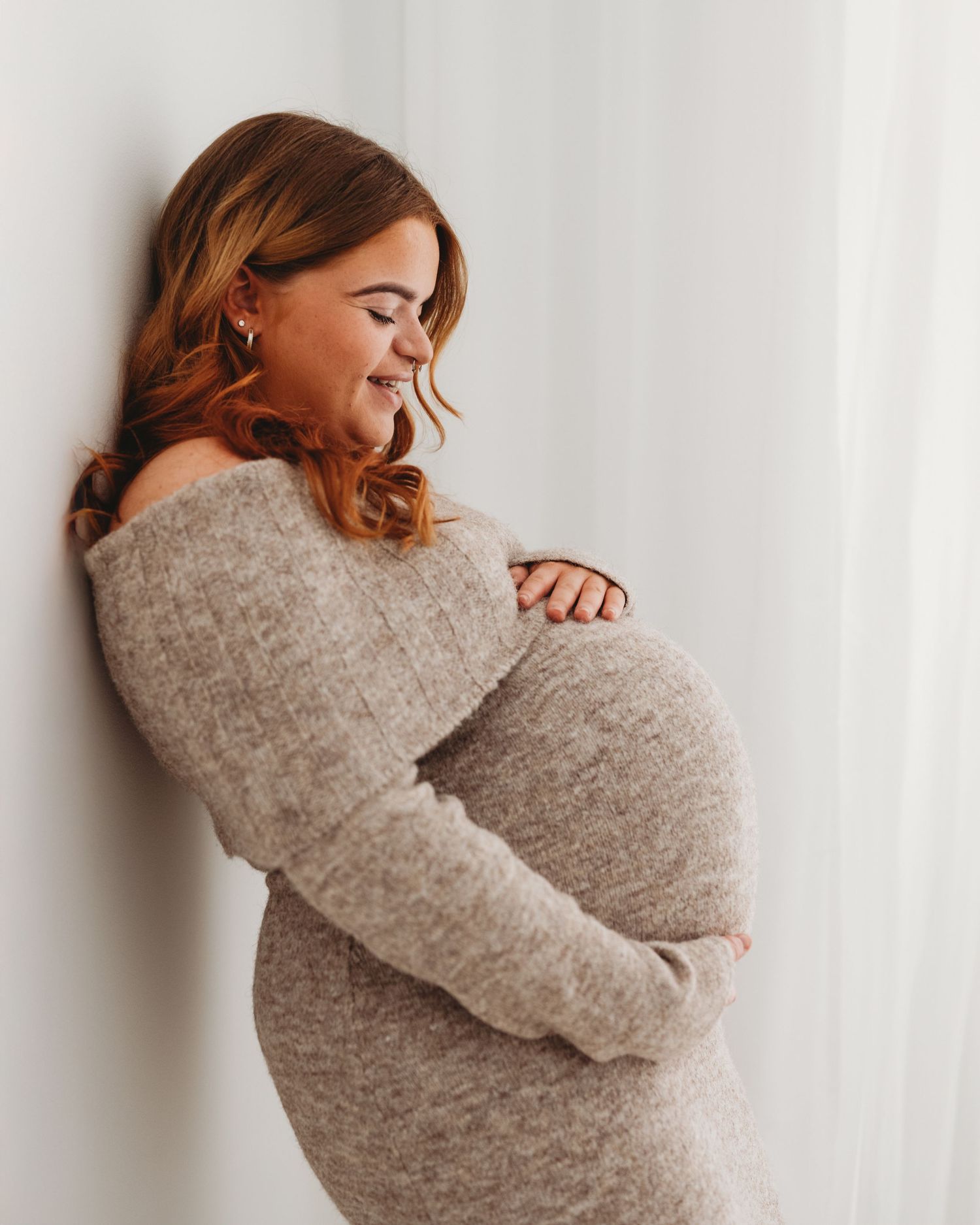 relaxed pregnant mother is captured naturally smiling, leaning against a wall in Faye McRae's all-white Ayrshire photography studio
